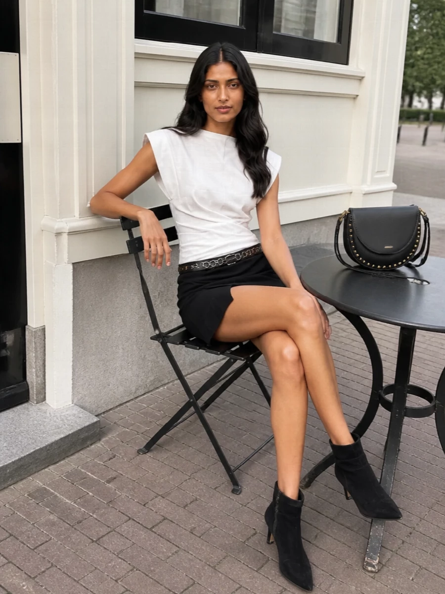 A sharp white sculpted top plays against the black wrap mini skirt for a clean, Paris-meets-street mood. Pointed suede ankle boots and a studded saddle bag lean it into after-dark territory, while the chain belt adds just enough hardware to keep the black-on-black elements interesting and intentional. The result feels leggy, graphic and ready for warm city evenings without ever drifting into fuss.
