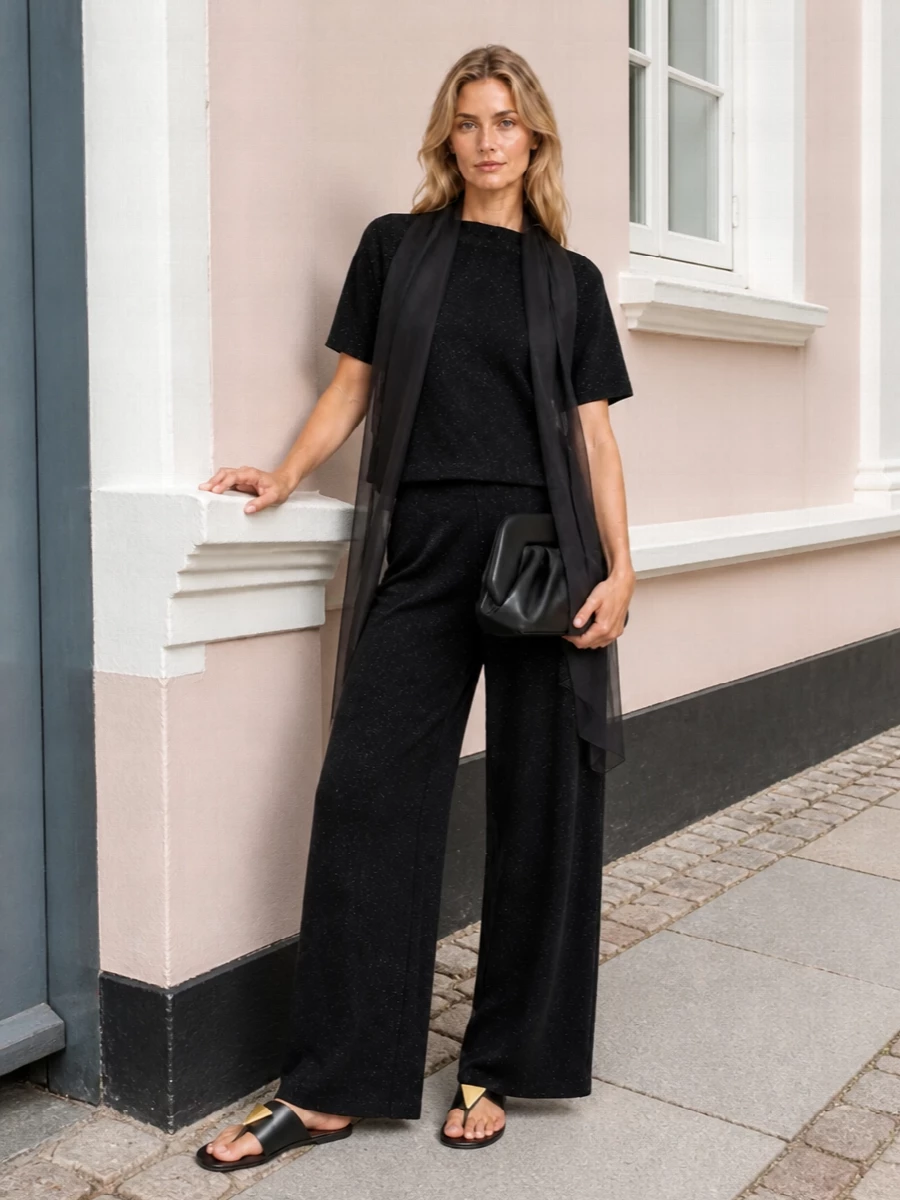 A head-to-toe dark palette lets the knit shimmer speak, paired with fluid wide-leg trousers for a soft, elongated line. Graphic black slides and a structured clutch keep the mood polished yet unfussy, while a sheer charcoal scarf adds movement and a whisper of texture. It is an easy day-to-dinner ensemble that feels quietly confident and modern.