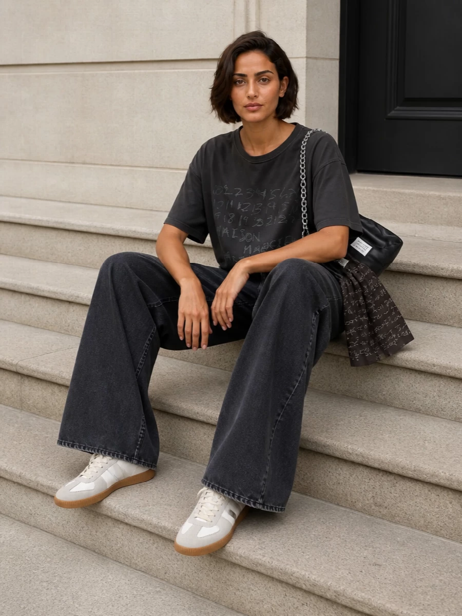A washed charcoal numbers tee sits against exaggerated wide-leg charcoal denim for a clean, elongated streetwear silhouette. Iconic white-and-gum sneakers lighten the base, while a padded black chain bag and matching numbers scarf lock in a cohesive Margiela graphic story. The result is relaxed yet intentional, all about proportion, texture and quiet branding.