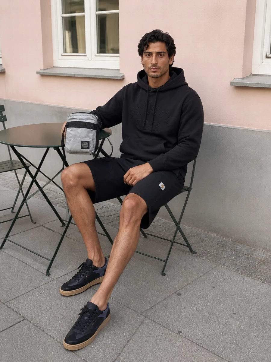 Carhartt cargo shorts meet a washed black Stüssy hoodie for a clean, skate-adjacent street uniform in deep charcoal tones. Black gum-sole Axel Arigato sneakers keep it low-profile but sharp, while the Stone Island crossbody introduces a small hit of tech texture and structure. The result is relaxed and utilitarian, ready for city days that move from daylight to late-night without a change.