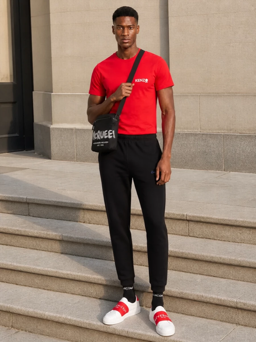 A vivid red Kenzo tee sits against clean black tapered track pants for a sharp, sport-led base. White slip-on sneakers with a red logo band amplify the color story while keeping the silhouette sleek. A compact black crossbody and subtle logo socks finish it with just enough graphic edge for city days and travel.