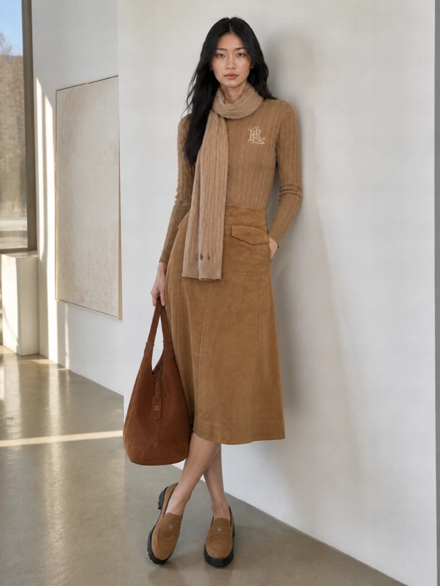 Camel on camel feels intentional here, with the cable-knit sweater and corduroy midi skirt creating a warm, tonal column that is quietly refined rather than fussy. Chunky-sole suede loafers keep the mood grounded and modern, while a cognac suede tote and slim camel scarf pull the textures together into a polished, heritage-leaning outfit for city days or country weekends.