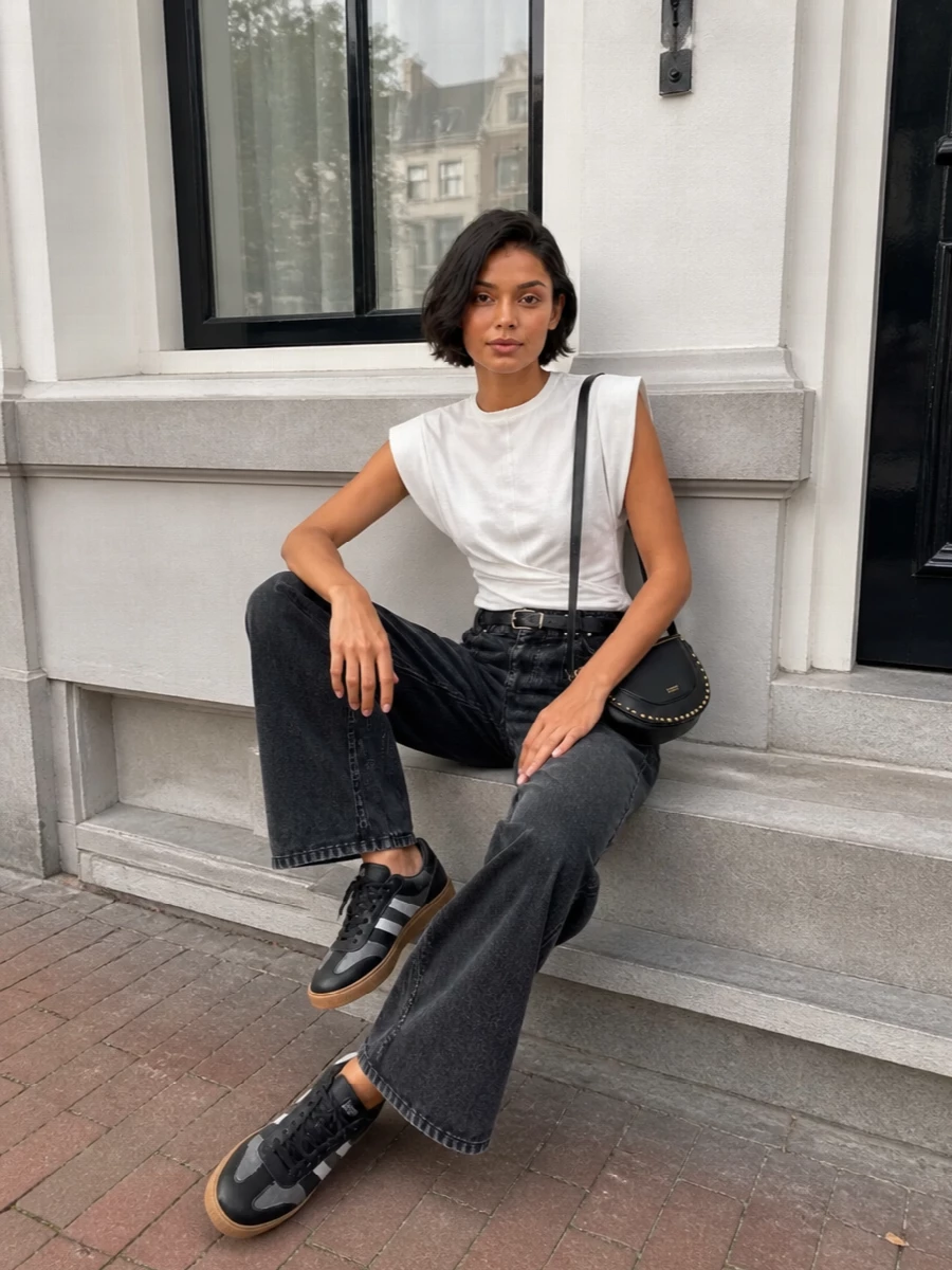 High-rise charcoal pleats meet a sculpted white top for a clean, graphic play on volume and restraint. Streamlined sneakers and a slim black belt keep it streetwise yet precise, while the compact saddle bag adds a subtle rock-boho note without disturbing the long leg line.