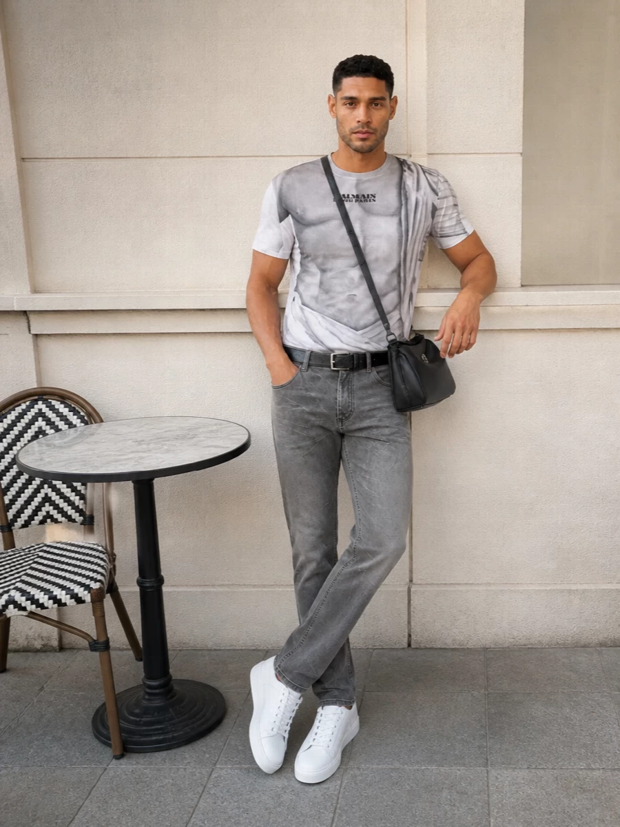 The marble-torso tee sits on washed-grey denim in a clean straight cut, so the whole outfit reads sharp and graphic rather than busy. Soft white sneakers keep the energy relaxed but precise, while a structured black crossbody and slim textured belt add just enough polish for city evenings or gallery days. It is a monochrome, tailored streetwear combination that lets the Balmain artwork stay firmly in the spotlight.