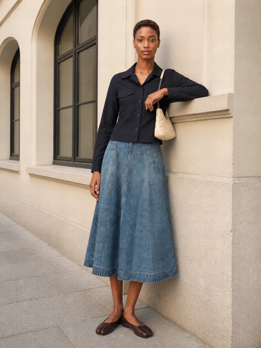 A mid-wash A-line skirt meets a sharply cut navy shirt for a clean, modern take on denim that still feels discreetly polished. Chocolate split-toe flats and a butter-soft crescent bag keep the palette muted and refined, adding just enough interest without disturbing the quiet balance.
