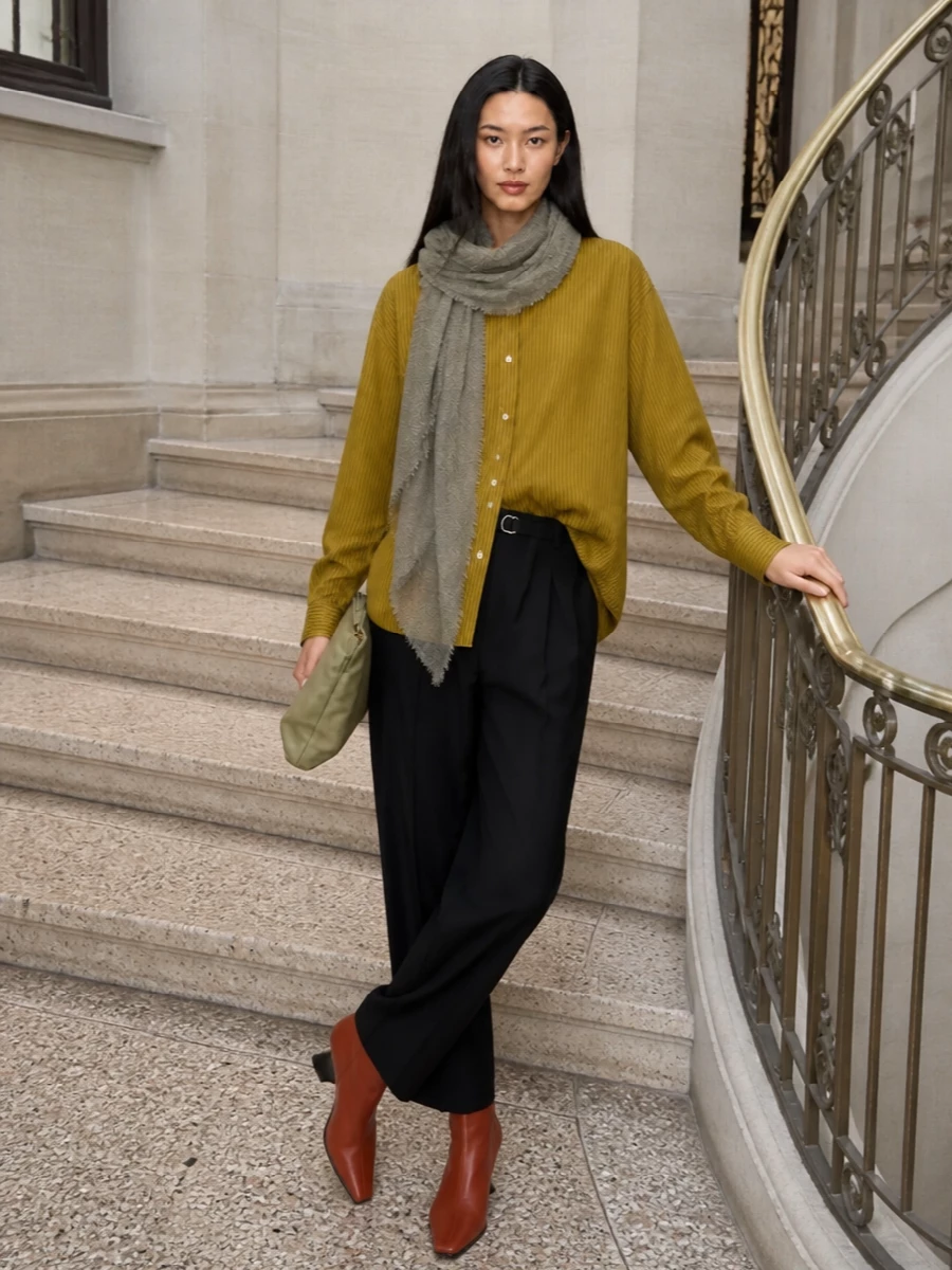 An oversized mustard stripe shirt falls over fluid black pleated trousers for a quietly assertive, elongated line. Rust leather ankle boots introduce a sculptural hit of color, while a sage-olive clutch and washed grey scarf keep the palette moody and intelligent. The result is polished and relaxed at once, ideal for days that move from desk to dinner without a change of attitude.