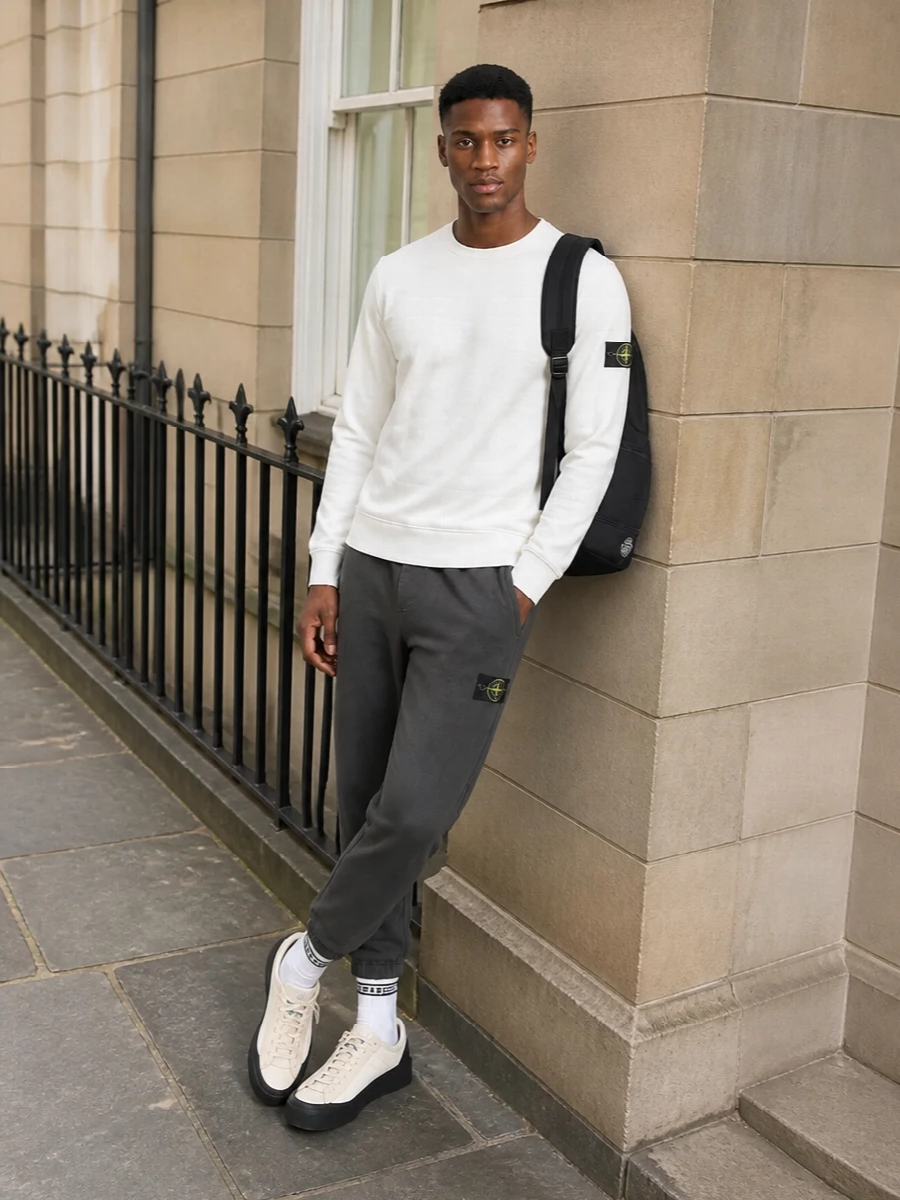 Optic white up top and washed charcoal below keep the palette tight and graphic, with each Stone Island badge reading as a deliberate detail rather than a shout. The cream and black sneakers echo that light‑meets‑dark balance, while the black technical backpack and striped socks ground everything firmly in a clean, urban sport mood that works from airport runs to city days.