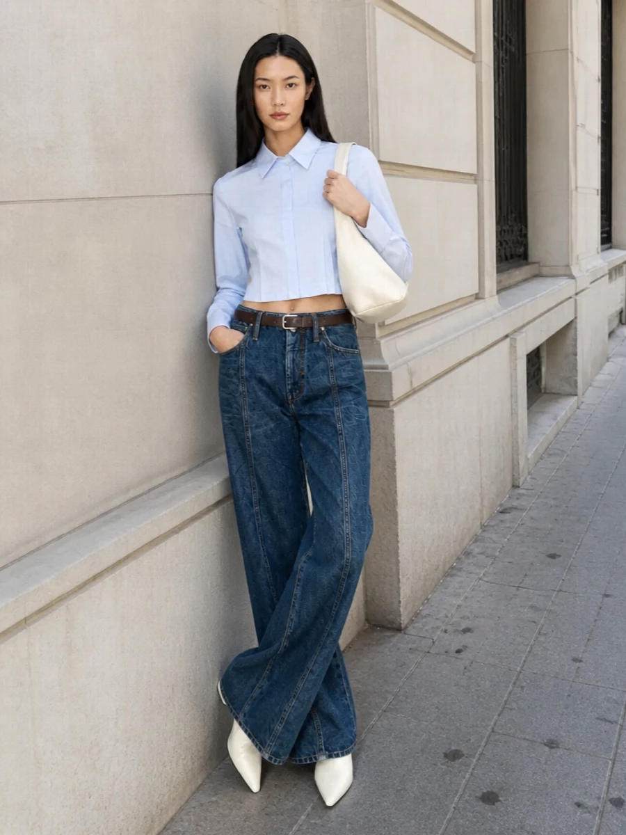 Architectural wide-leg jeans are grounded with a cropped sky-blue Sportmax shirt, giving a clean, elongated upper half that offsets the volume below. Soft white pointed flats and a sculpted ivory shoulder bag keep the palette light and refined, while a slim brown belt subtly echoes the denim’s topstitching for a cohesive, quietly luxurious SS look.