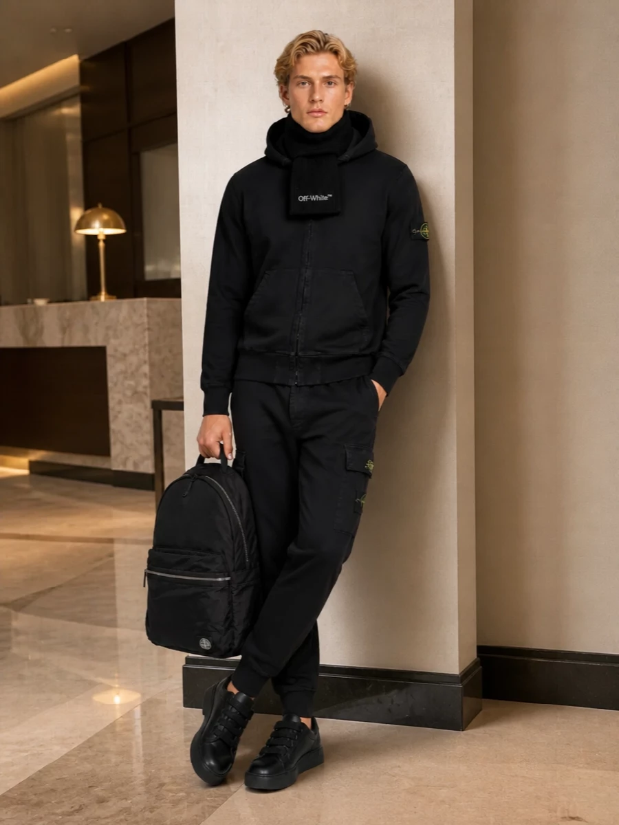 Washed black on black keeps this Stone Island hoodie and cargo jogger combination tight and utilitarian, with technical details doing the quiet work. A chunky all-black sneaker grounds the volume and adds polish, while the nylon backpack and ribbed scarf lean into a city-ready, sport-informed winter uniform.