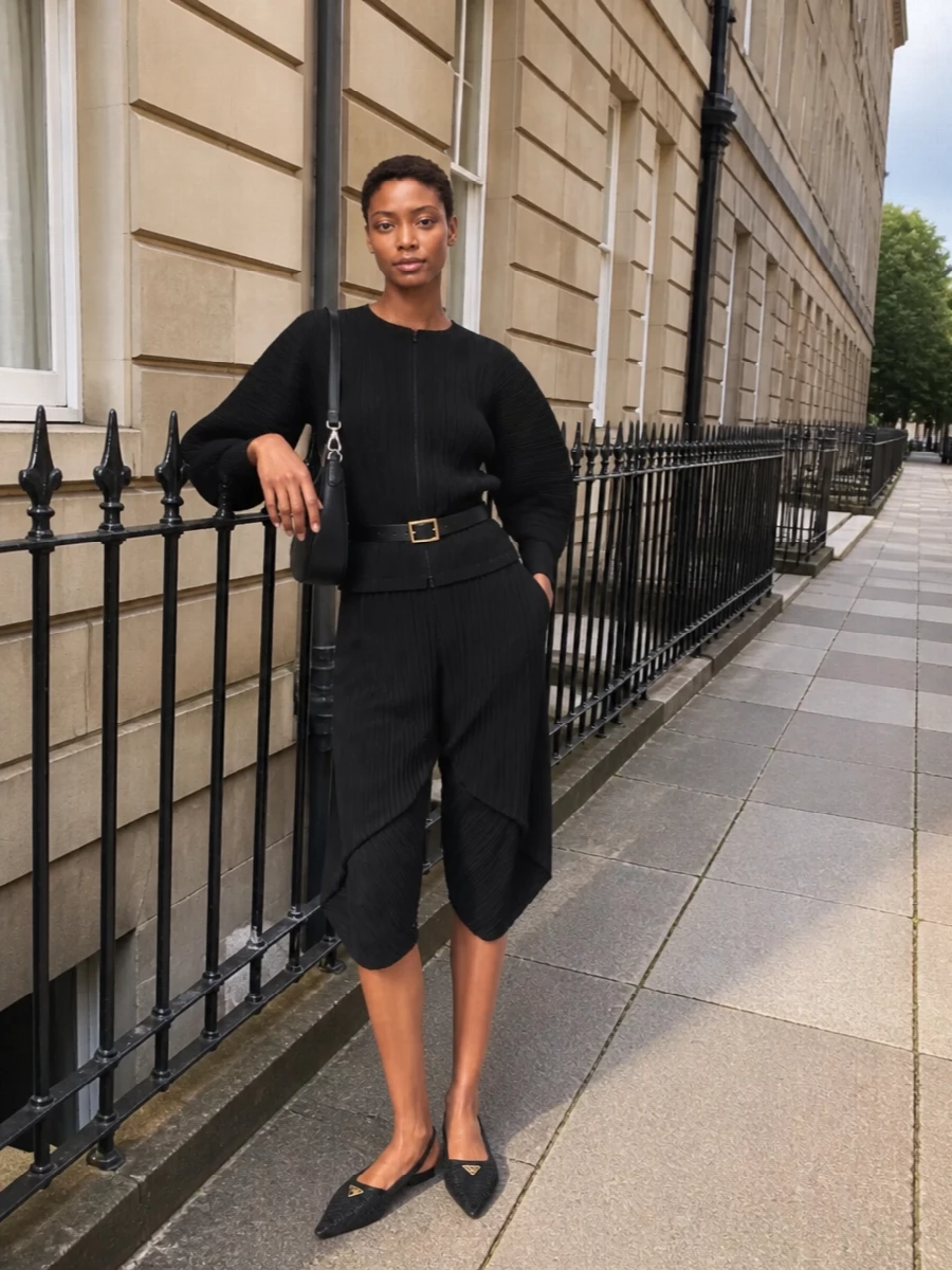 An all-black Pleats Please set keeps the focus on sculptural pleating, with the cocooned jacket-top and asymmetric short reading as a single, fluid column. Pointed Prada flats and a crescent shoulder bag keep everything low to the ground yet sharply finished, so the movement of the fabric and the architecture of the silhouette do all the work. A slim black belt is there if you want to cinch the waist and dial the structure up a notch without losing ease.