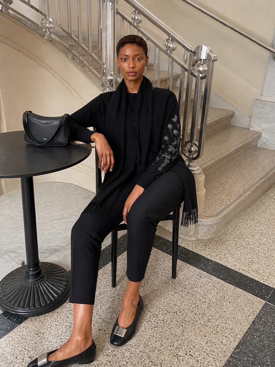 A black-on-black palette lets the sweatshirt’s silver sleeve embellishment take focus, grounded by slim cropped trousers that keep the line sharp but relaxed. Soft, directional ballet flats and a structured cross-body keep the mood quietly polished for city days while staying easy to move in. The black scarf adds a subtle, tactile layer without disturbing the minimalist balance.
