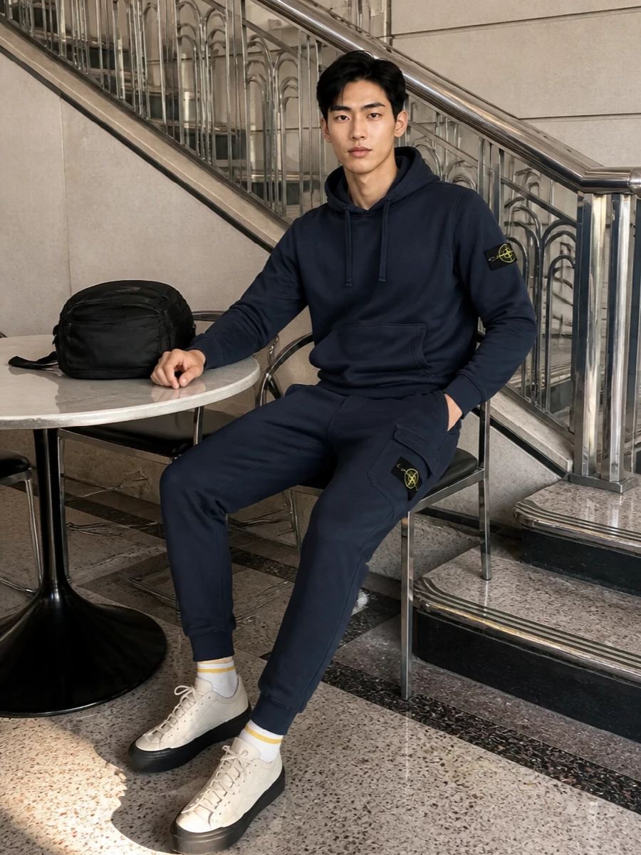 A navy Stone Island hoodie sits over tonal cargo joggers for a clean, badge-to-badge street set that feels put together without trying. Neutral Stone Island runners and a black technical backpack keep the lines streamlined and practical, while striped socks quietly echo the brand’s yellow detailing. It is an easy travel or city-uniform formula you can repeat all season with just a tee swap underneath.