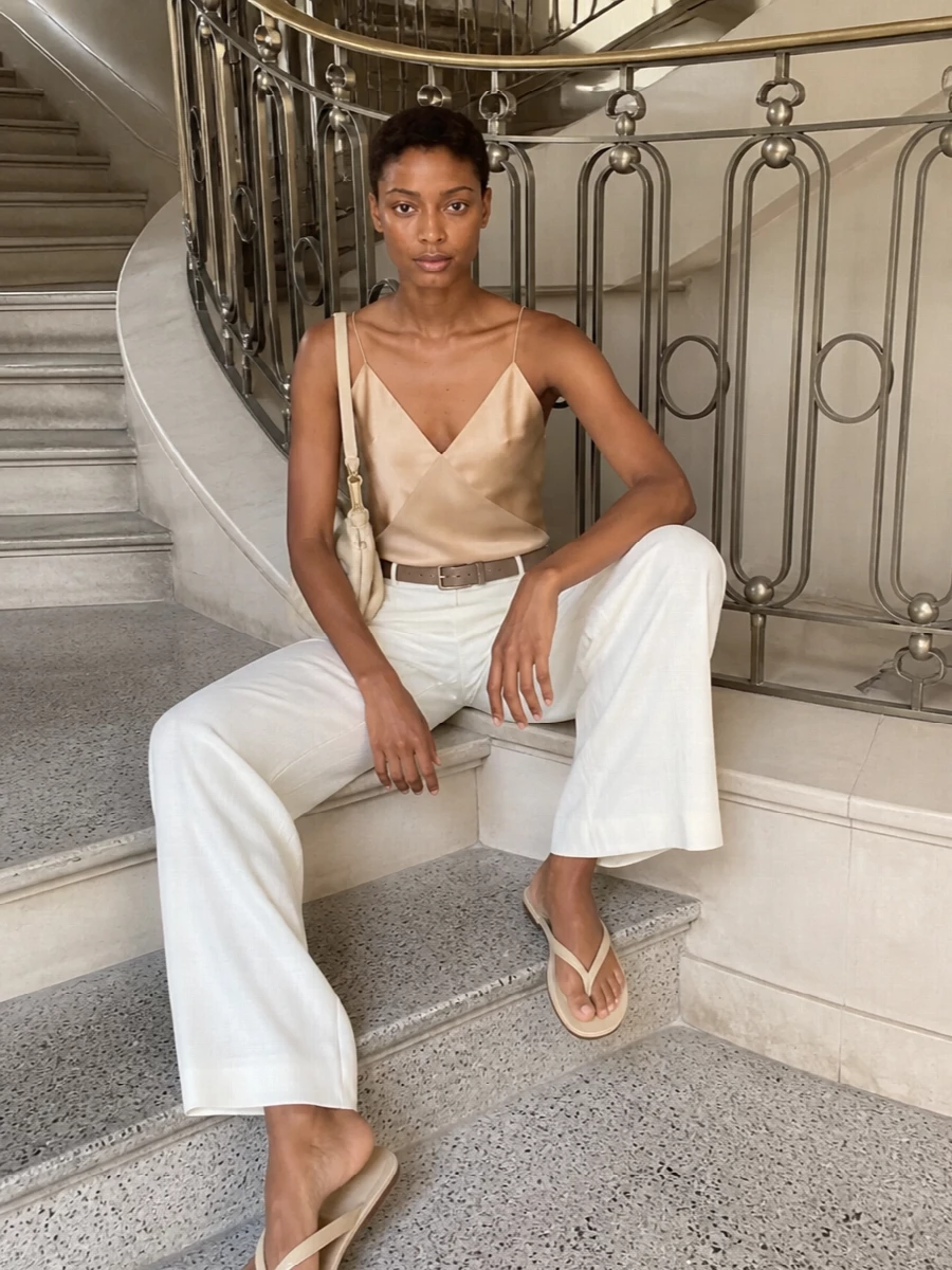 A soft champagne camisole and fluid ivory trousers create a long, quiet column of light neutrals. Buttercream sandals and a pleated shoulder bag keep the mood relaxed yet refined, while a slim taupe belt offers just enough structure without disturbing the ease.