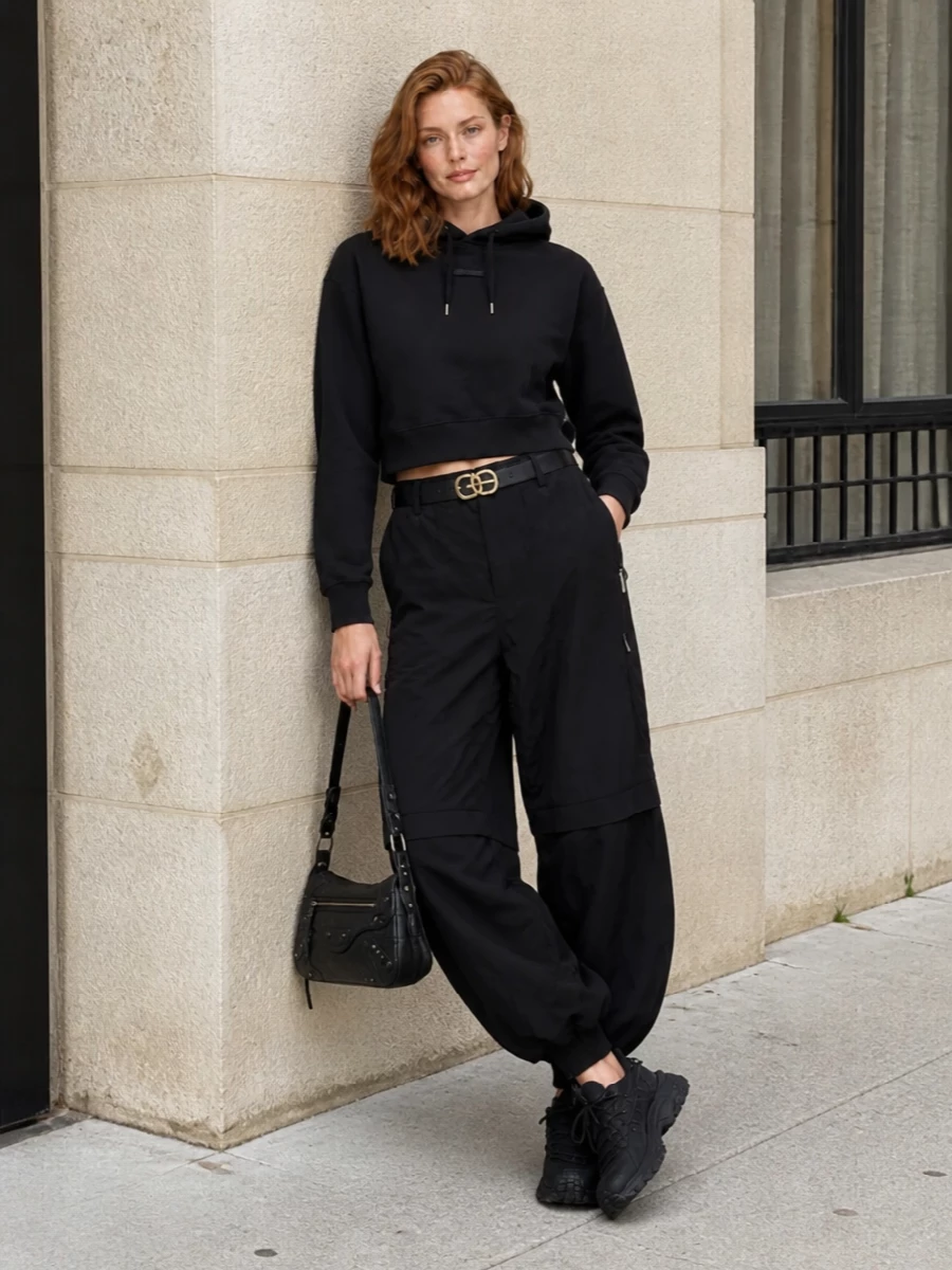 This look builds a monochrome, sculptural streetwear silhouette: the cropped black Jacquemus hoodie sits neatly over the dramatically ballooned track pants, emphasizing the volume through the leg while keeping the torso clean. Chunky black Moncler sneakers anchor the proportions with a technical edge, and a compact black Balenciaga shoulder bag plus a slim logo belt sharpen the finish, delivering a polished yet easy FW city uniform.