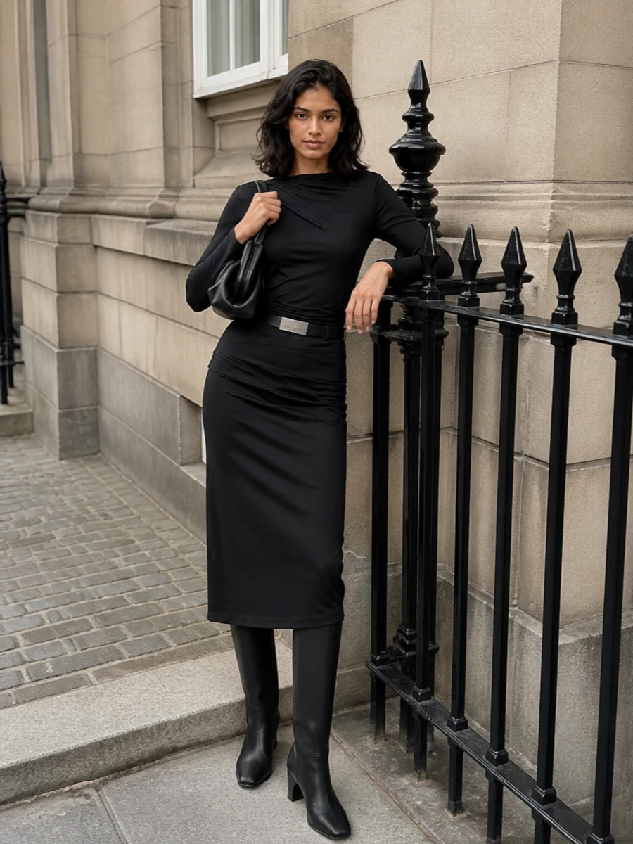 An all-black, body-skimming column keeps the asymmetric midi at the center, with the draped jersey top subtly mirroring its wrap for a clean, directional line. Sculptural knee-high boots extend the silhouette, while a compact pleated shoulder bag and a precise slim belt add just enough structure for gallery evenings or late cocktails without cluttering the minimal mood.