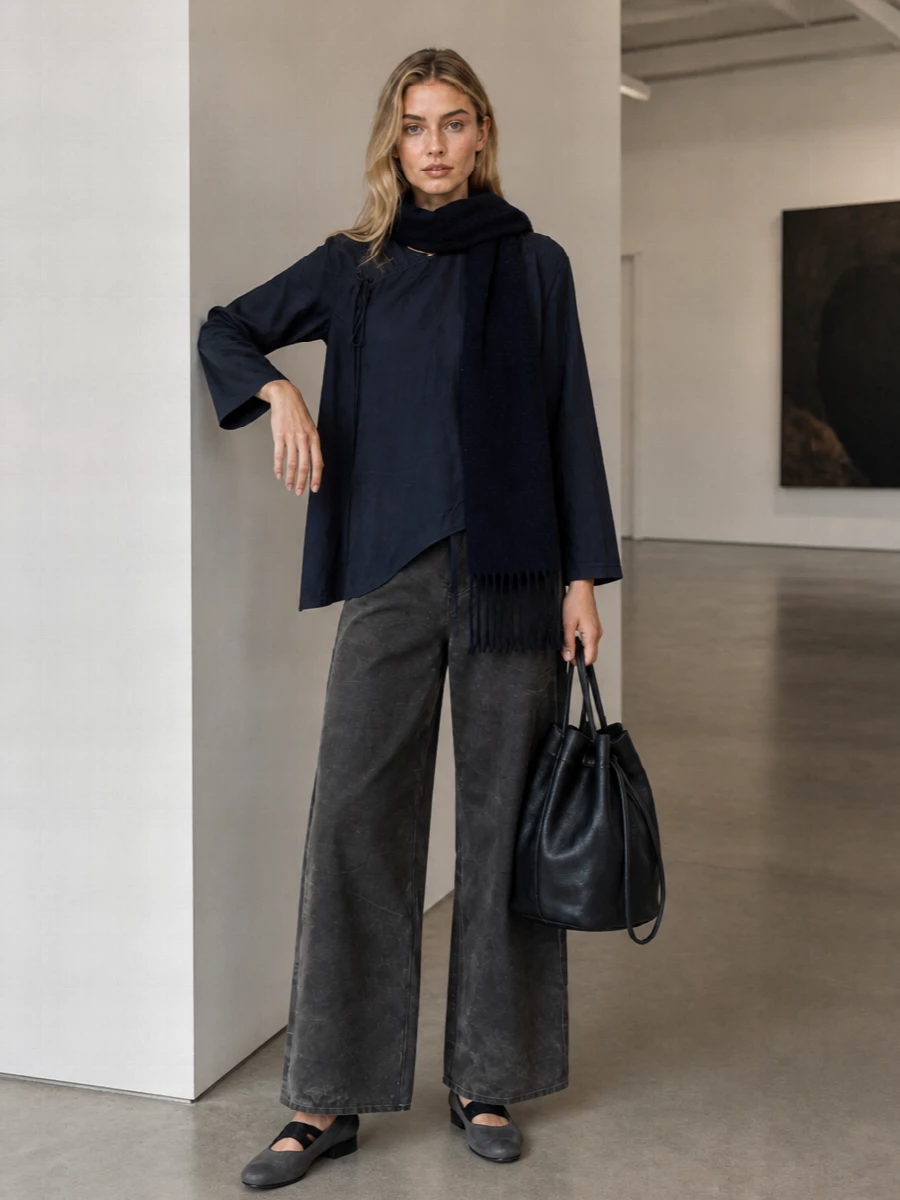 Deep navy and washed charcoal sit together in a quietly tonal column, the asymmetric tunic skimming over full, architectural trousers. Charcoal block-heel ballerinas and a soft black bucket bag keep the mood grounded and textural rather than delicate. A navy scarf echoes the top’s shade, pulling the palette into a calm, sculptural whole suited to thoughtful city days or gallery evenings.