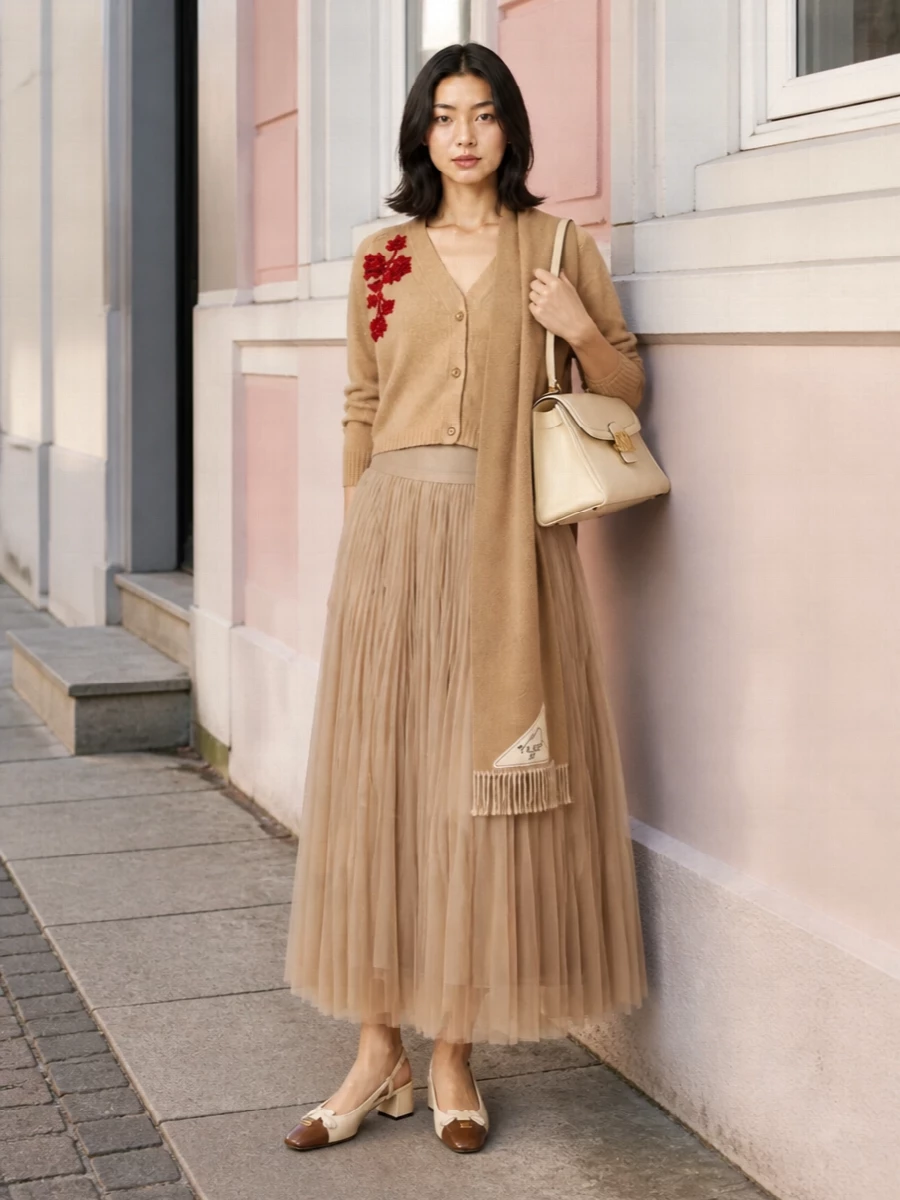 A cropped camel cardigan with crimson embroidery tops a feather-light pleated maxi, all in warm neutrals that feel soft yet intentional. Cream and chestnut slingbacks and a structured buttercream bag sharpen the romance just enough for polished dinners or gallery evenings. The long, fluid skirt against the neat knit keeps the silhouette graceful and quietly elongating.