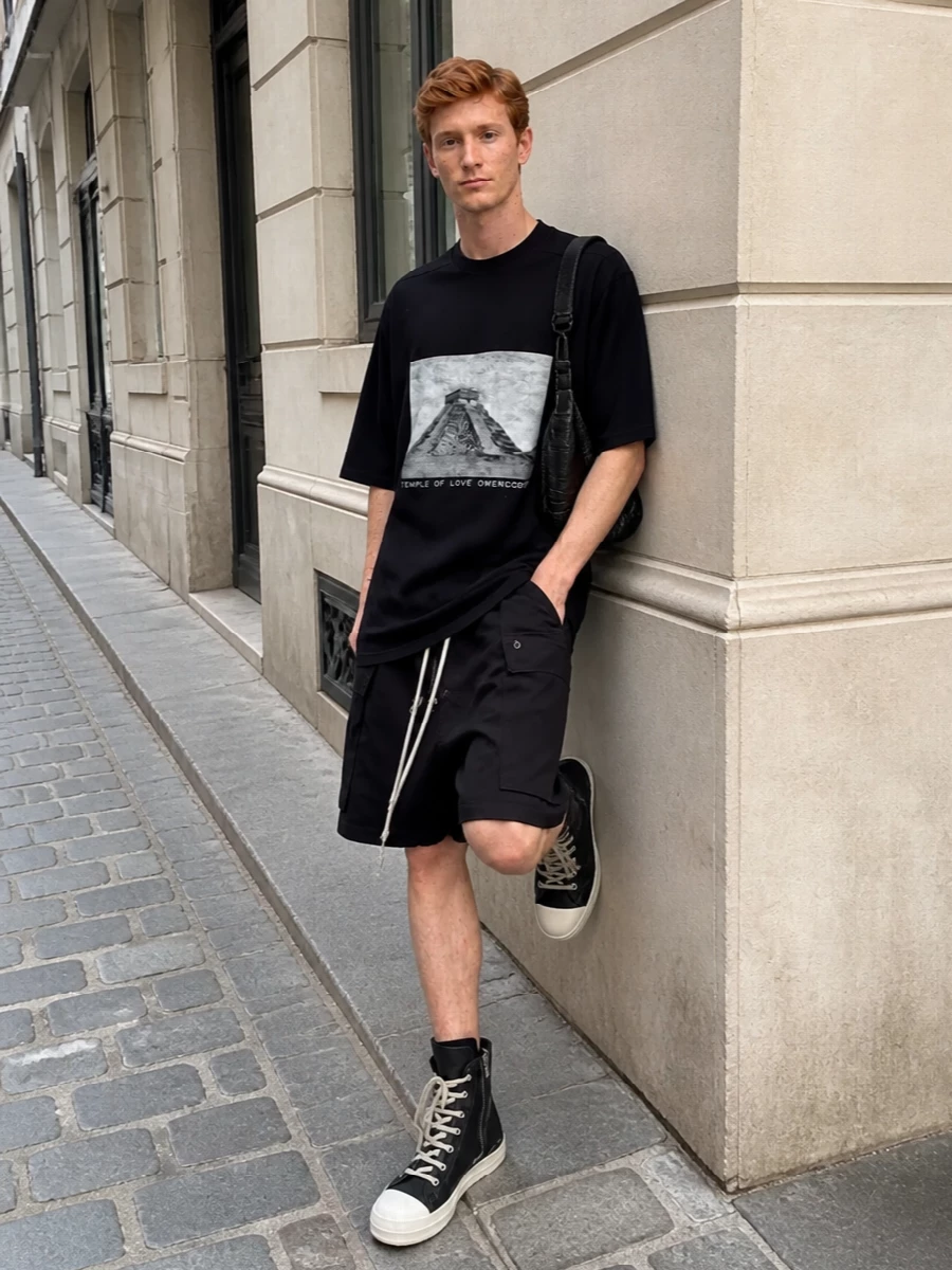 The look leans fully into Rick’s street‑utility universe: a longline black temple graphic tee dropping over black DRKSHDW cargo shorts with a relaxed, low‑slung crotch and exposed drawcords. Chunky black hi‑top sneakers with contrast foxing extend the stacked, architectural line, while a compact black leather shoulder bag introduces just enough hardware and polish to keep the outfit sharp for summer city wear. The result is slouchy, monochrome and directional, but still easy to live in every day.