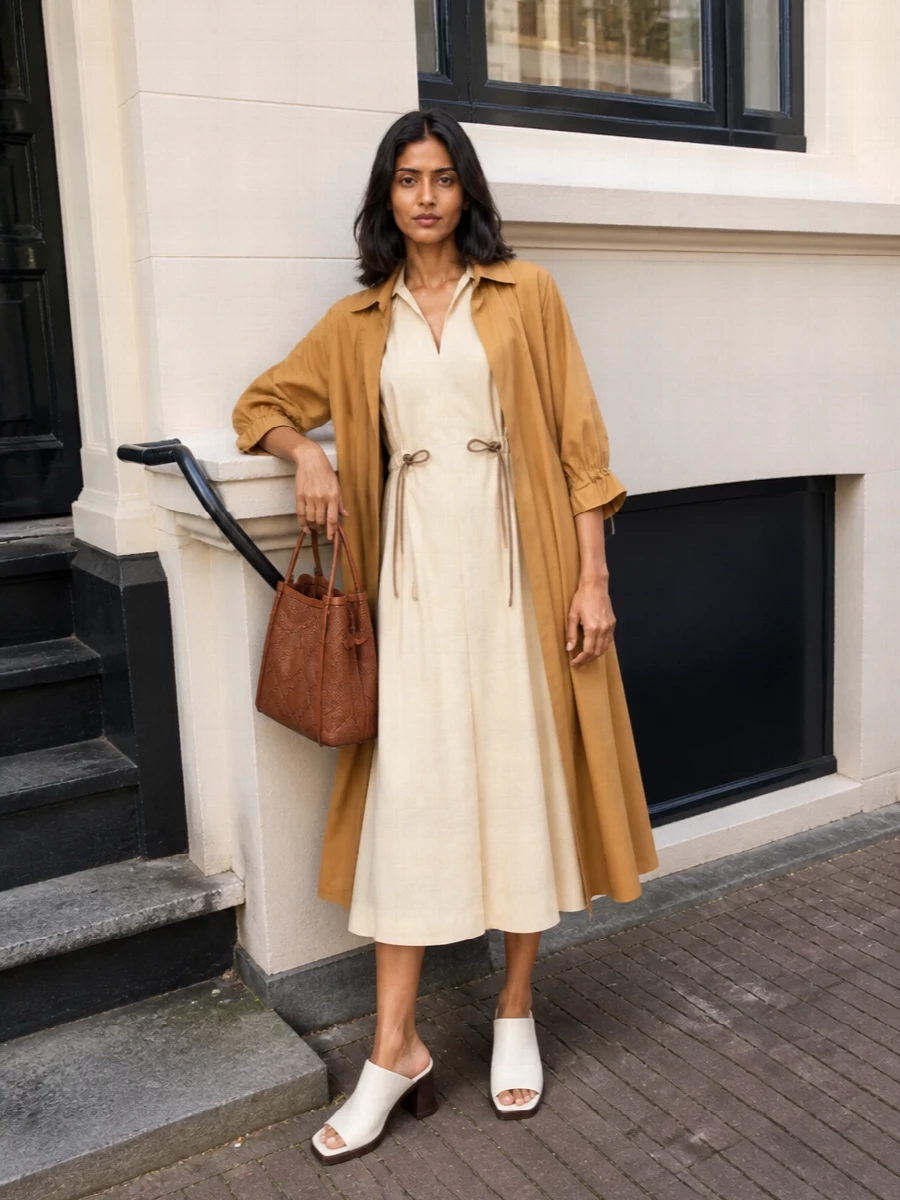 I am styling the trench with a shirt dress rather than tailoring separates, so the coat’s belt and A-line flare can read as the main structure over a single clean column. The light beige dress keeps things airy and softly defined, while ivory mules and a cognac woven bucket bag add just enough formality and texture for city days that can stretch into dinner. The result is a tonal neutral ensemble that feels composed, feminine and quietly confident without ever looking overworked.