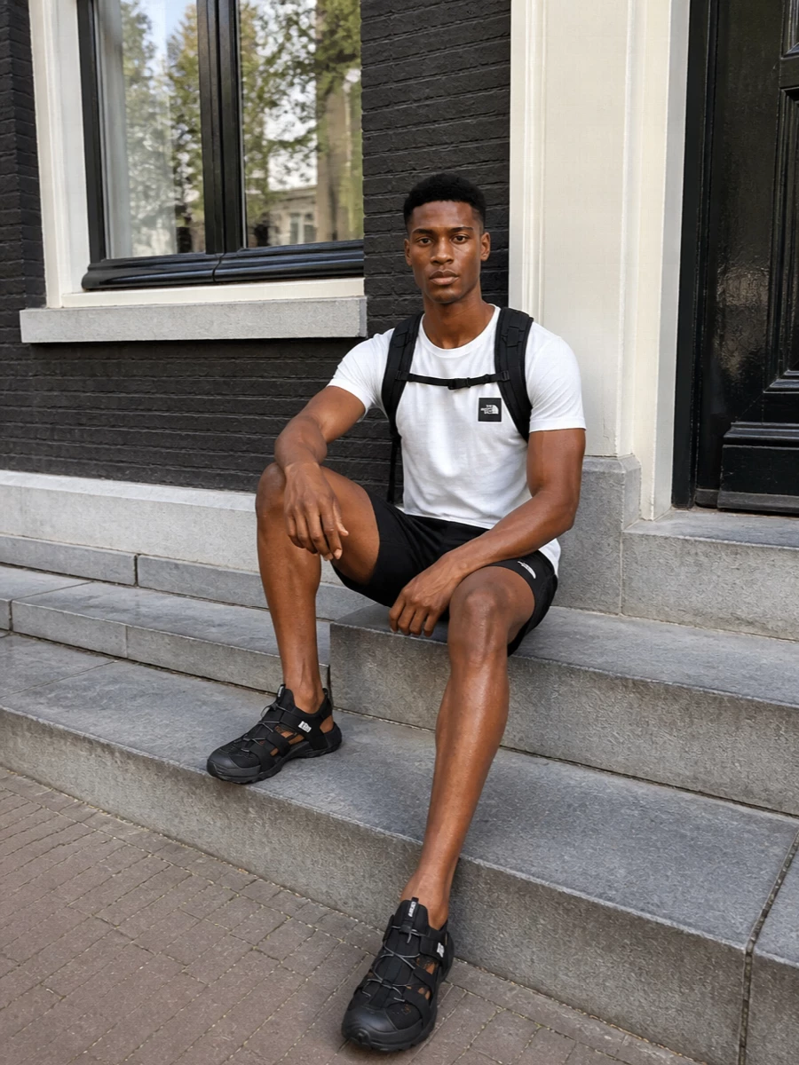 A crisp white logo tee sits against black technical shorts for a sharp, minimal take on trail-inspired streetwear. Closed-heel trekking sandals keep the line active and grounded, while the compact technical backpack completes the monochrome, movement-ready silhouette.