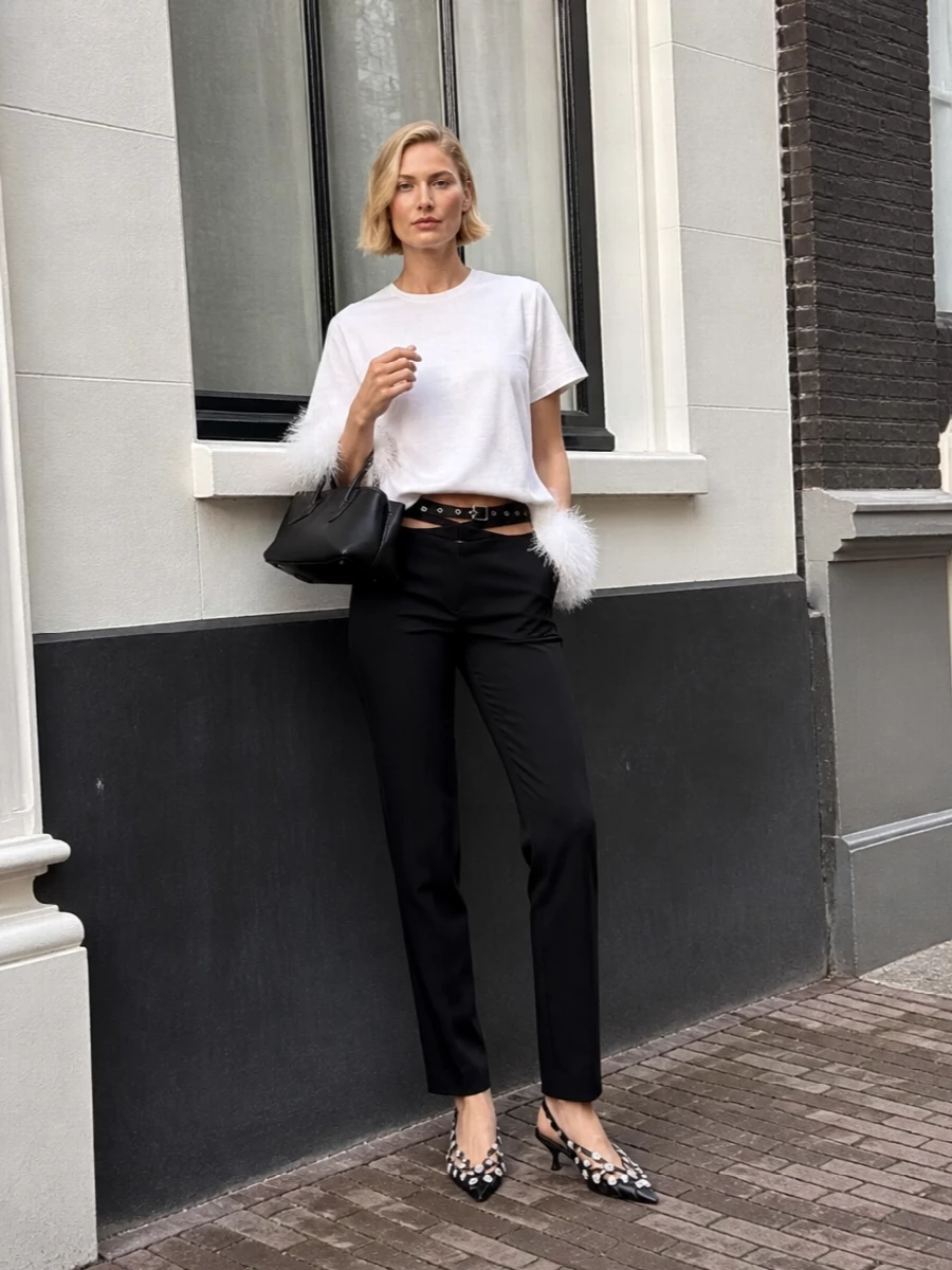 A feather-trimmed Attico tee becomes a party mini, paired with cut-out high-rise black trousers for a long, lean line and a hit of skin at the waist. Studded kitten-heel slingbacks and a sharp black mini bag keep everything graphic and nightlife-ready, while a slim eyelet belt ties the silver hardware together without distracting from the feathers.
