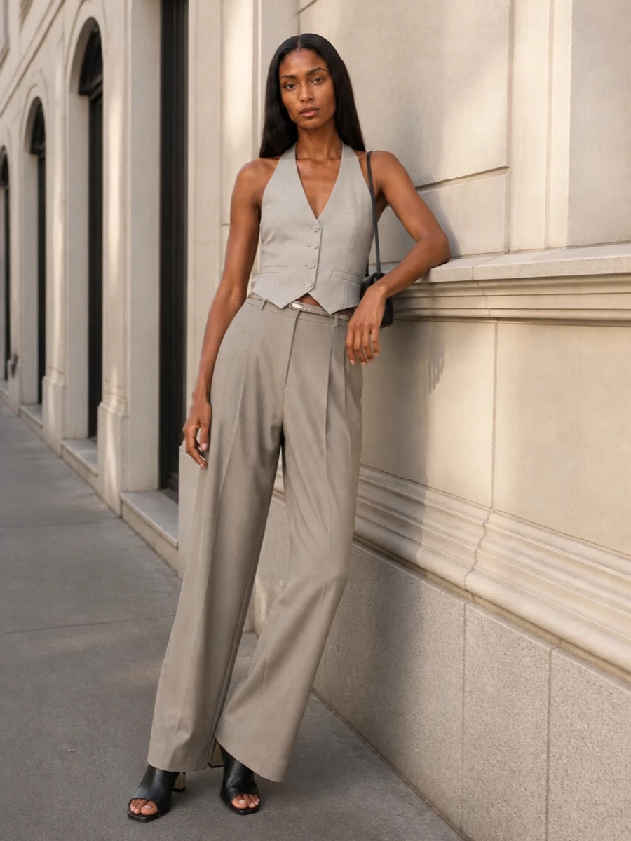 A quietly precise taupe-on-taupe column lets the halterneck waistcoat read as a modern tailored top, with wide-leg trousers elongating the line. Black sculptural mules and a slim shoulder bag introduce a crisp, graphic counterpoint, while a whisper of metallic at the waist keeps the palette soft and considered. The result is summer tailoring that feels sharp yet relaxed enough for long city days that turn into dinner.