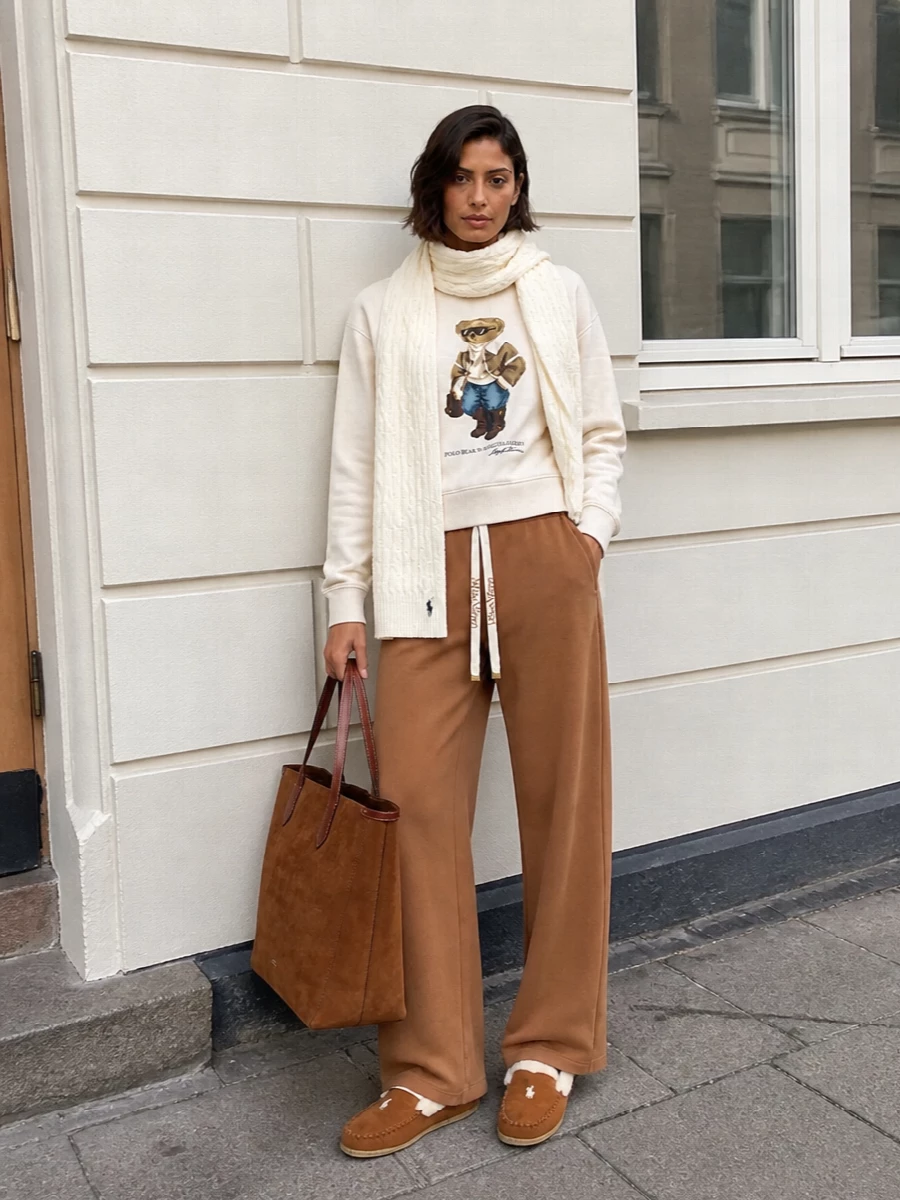 Cream, camel and caramel settle into an easy Polo Bear story that feels polished enough for coffee runs yet soft enough for the sofa. The relaxed sweatshirt pairs with fluid wide-leg lounge pants so the volume reads intentional rather than bulky, while the suede tote and moccasin slippers tie everything together in warm, collegiate neutrals.