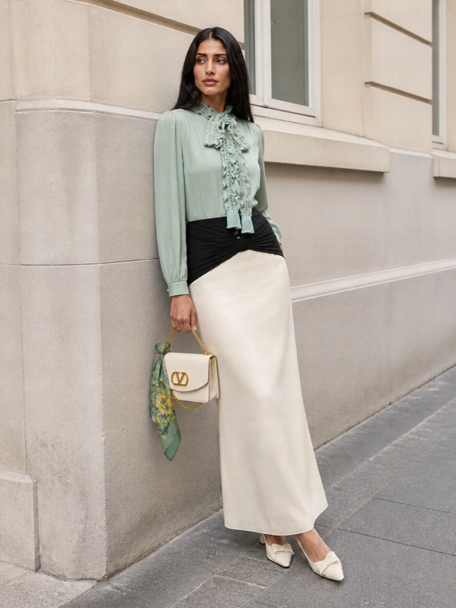 This look pairs the ruffled sage Valentino blouse with a cream-and-black fluid maxi skirt for a romantic, elongated silhouette that feels impeccably formal yet effortless. Buttercream accessories in the low-heel pumps and structured flap bag keep the palette soft and luminous, while touches of gold quietly echo the blouse’s buttons for a cohesive, quiet-luxury finish. A sage-trimmed floral scarf adds just a whisper of print and colour depth, enhancing the blouse rather than competing with it.