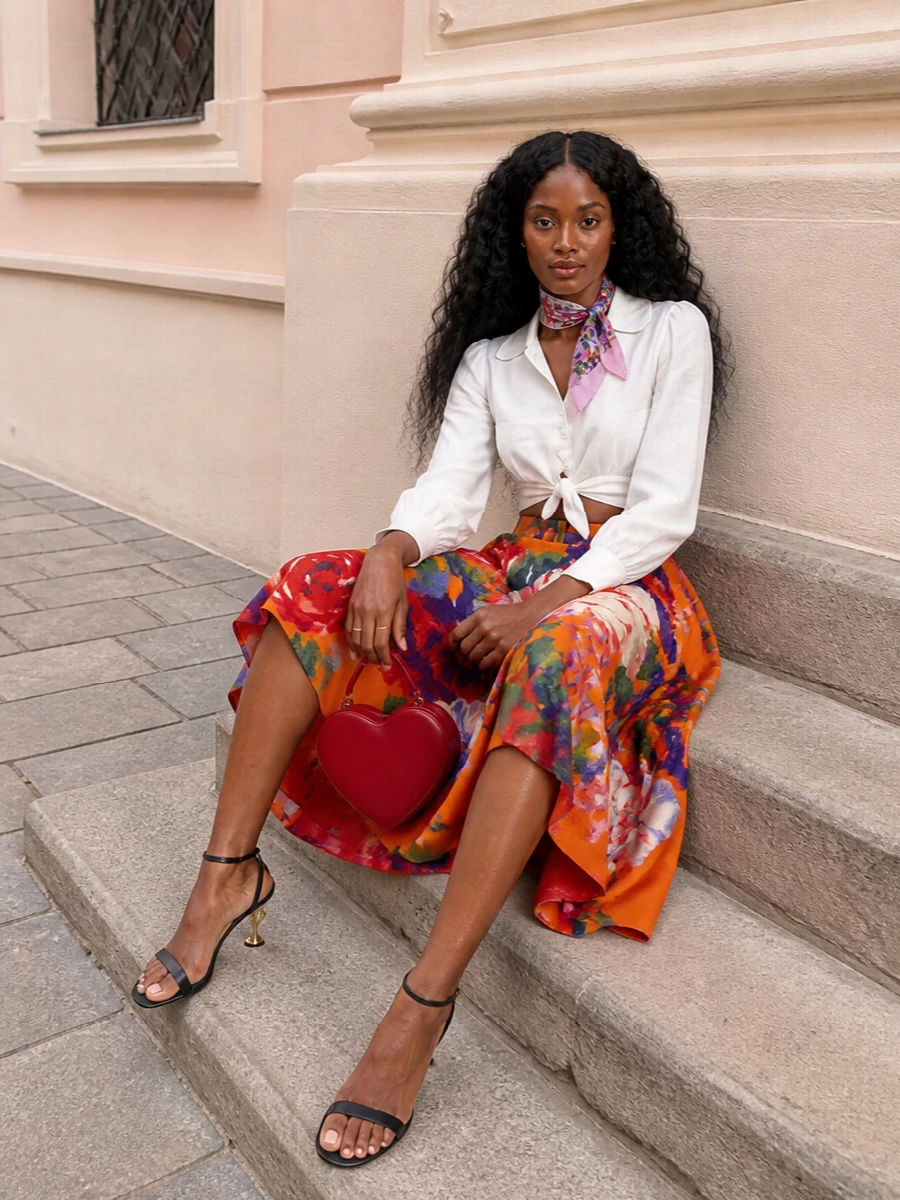 A vivid floral Moschino skirt takes center stage, tempered by a clean white tie-front blouse that sketches out a neat, feminine waist. Black and gold sandals add polished structure, while the red heart mini bag and floral scarf nod to the garden-party palette in a focused, romantic way. The result is joyful, refined and ready for sunlit terraces or summer weddings where colour is the point.