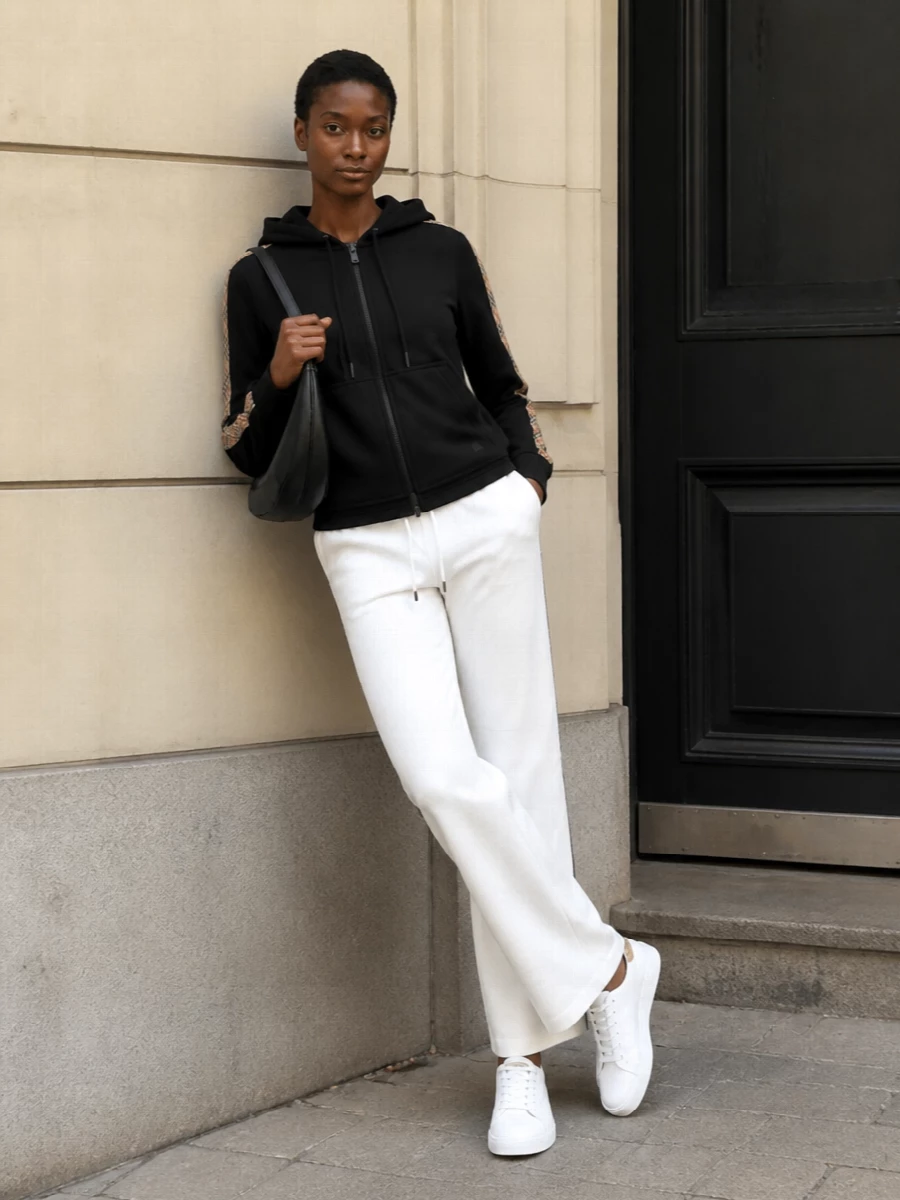 This look leans into elevated athleisure: the black Burberry zip hoodie is balanced by clean, soft-white straight-leg track pants for a streamlined, modern take on heritage sport. White leather sneakers with a whisper of check keep the palette fresh and cohesive, while the curved black shoulder bag adds a refined, urban finish that feels quietly luxurious rather than logo-heavy. It’s an easy, travel-ready uniform that still reads considered and polished.