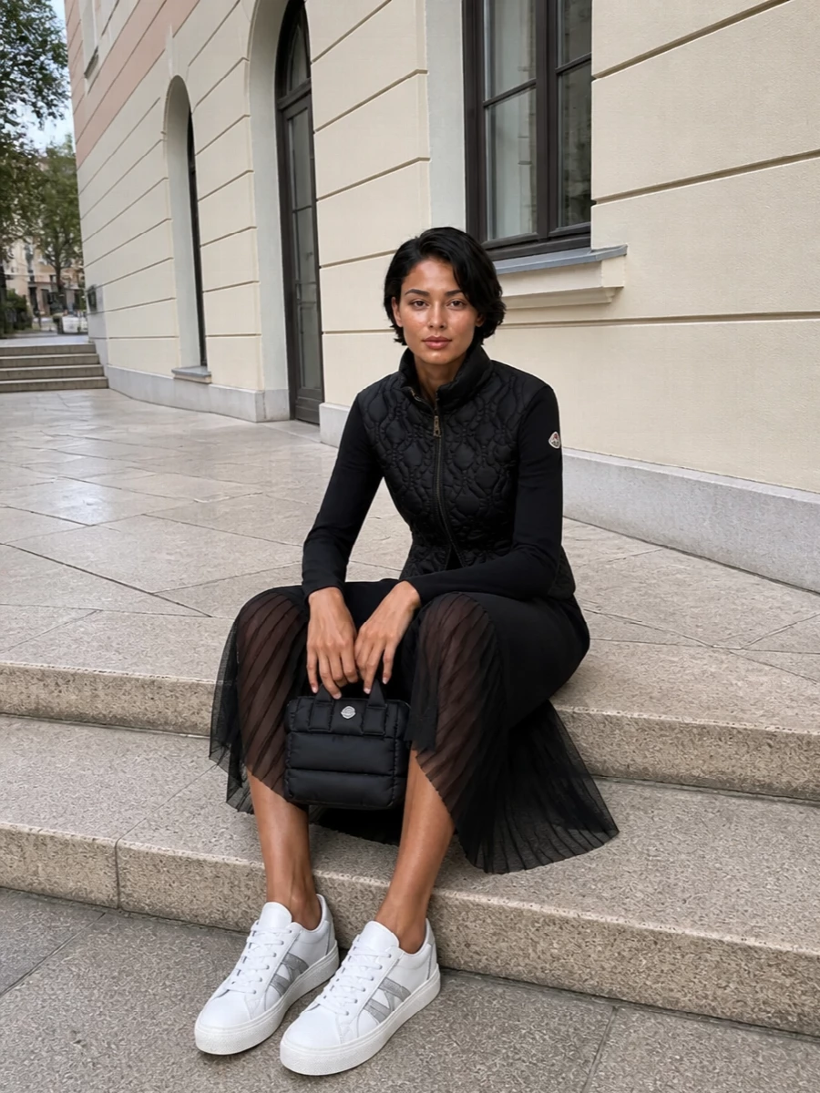 I chose the dress route, letting a single Moncler knit-backed puffer jacket top a simple black T-shirt midi to avoid over-layering padded separates. The monochrome column under the slim, quilted outerwear keeps everything quietly graphic and elongating, while white leather sneakers and a small padded tote pull it into an urban, sport-driven register. It feels like a refined city spin on après-ski, practical for cold days without losing that clean, minimalist edge.