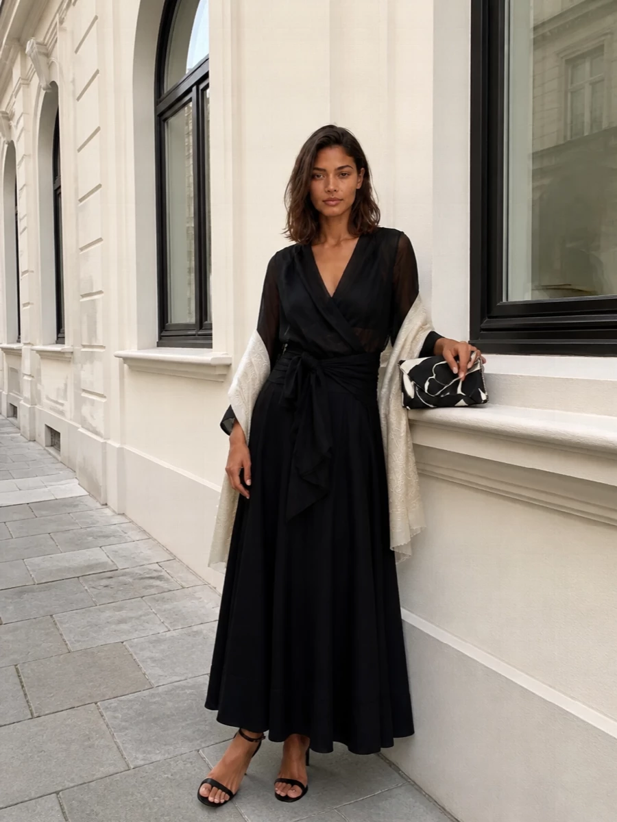A sheer black wrap blouse and a high-waisted flared maxi skirt create a long, liquid column that feels quietly powerful for evening. Barely-there black stilettos and a monochrome floral clutch keep the mood refined and modern. A tonal paisley stole adds just a hint of softness around the shoulders without disturbing the clean line.