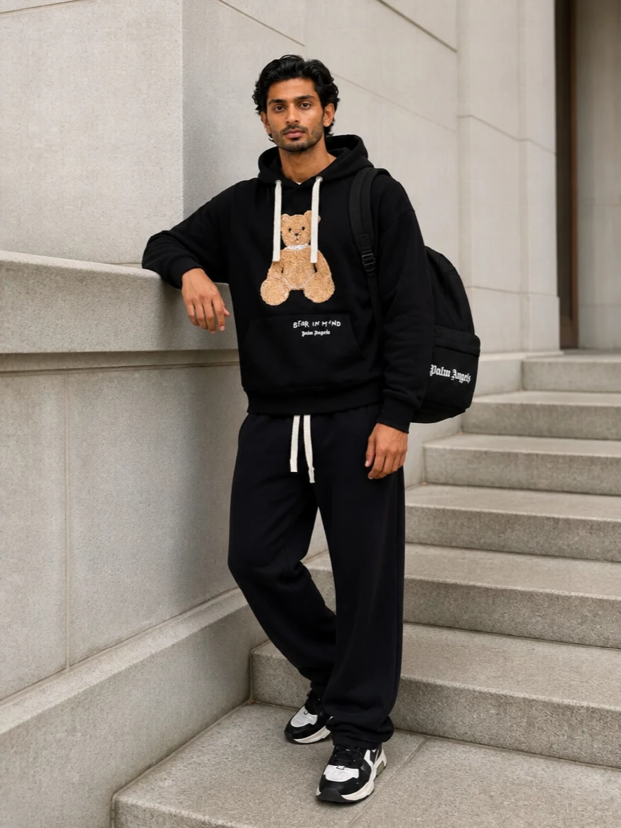 An oversized Palm Angels teddy hoodie sits over washed‑black relaxed track pants for a soft‑meets‑sport street uniform in a tight monochrome palette. Chunky Off‑White sneakers plug in subtle graphic detail and height without stealing focus, while a clean Palm Angels backpack keeps the branding consistent and the silhouette practical for city days. The result is an easy, confident off‑duty set that feels curated rather than casual.