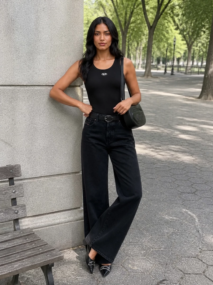 High-rise washed black denim and a tight ribbed black tank create a strong column of dark, minimalist streetwear energy. A sharp pointed flat and compact black shoulder bag add metal accents without softening the attitude. The slim black belt quietly cinches the waist so the wide leg reads intentional and architectural, not overwhelming.