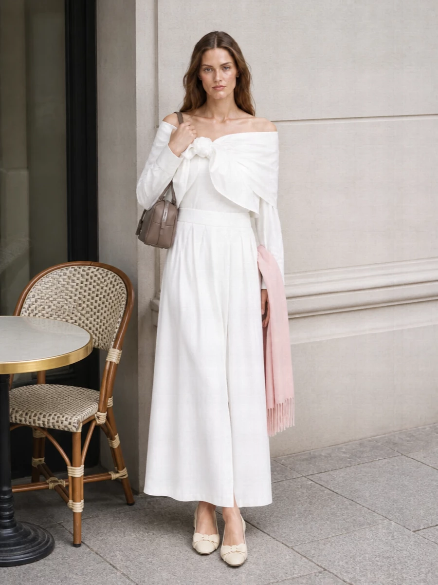 An off-the-shoulder bow-tied blouse pairs with a crisp white pleated maxi skirt to create a sculptural, monochrome column that feels poised yet soft. Cream block-heel pumps and a structured taupe mini box bag keep the palette refined and the silhouette elongated, while a blush scarf adds a whisper of colour that stays firmly within the quiet-luxury mood. Ideal for warm-weather dinners, terraces, or formal daytime occasions where you want romance with architectural clarity.