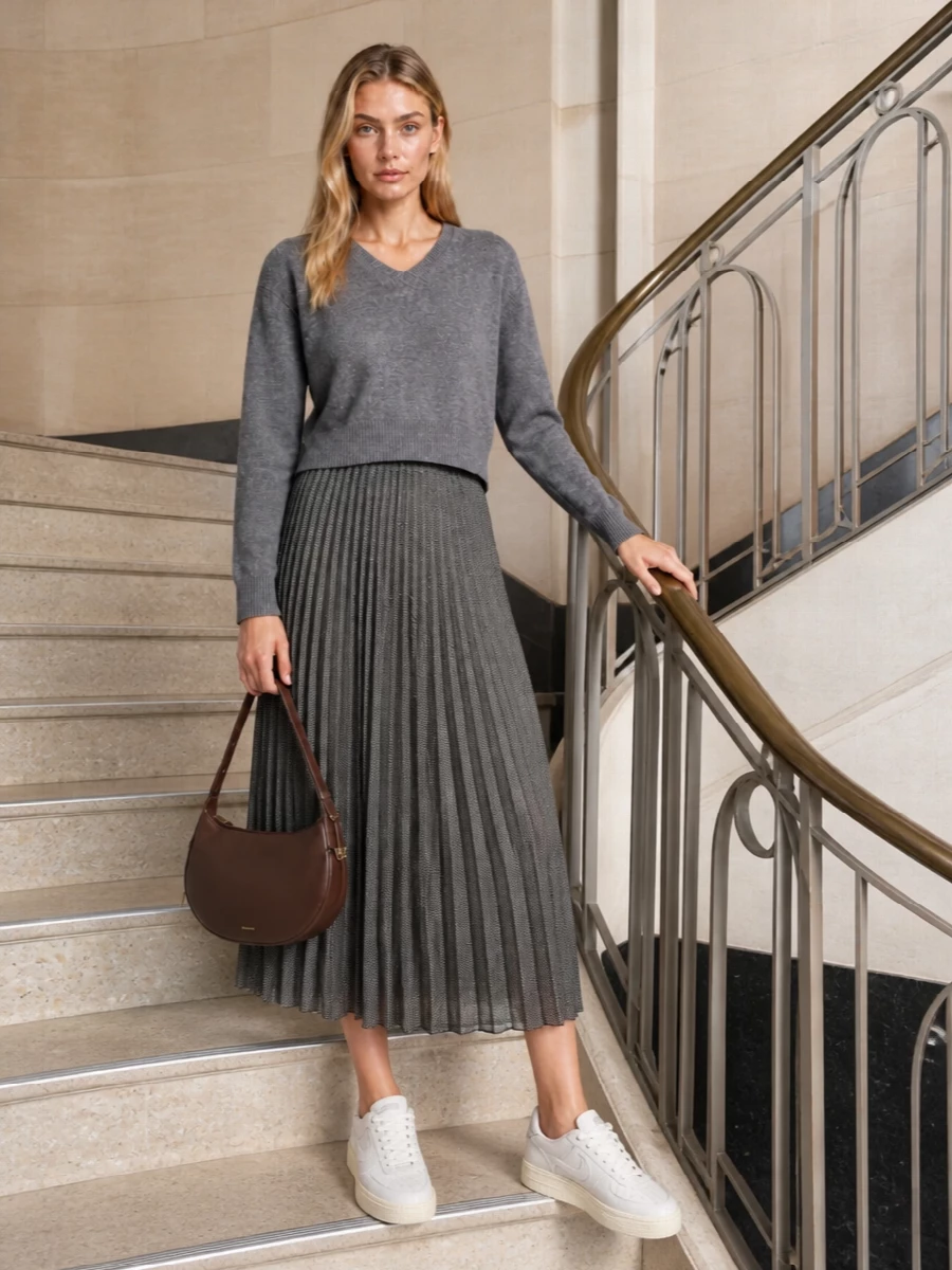 Heathered grey on soft charcoal checks keeps the palette hushed while the pleated midi adds movement and a slightly more dressed feel than denim. Clean off-white sneakers keep it easy for day, and the curved chocolate shoulder bag warms everything up without breaking the minimalist line.