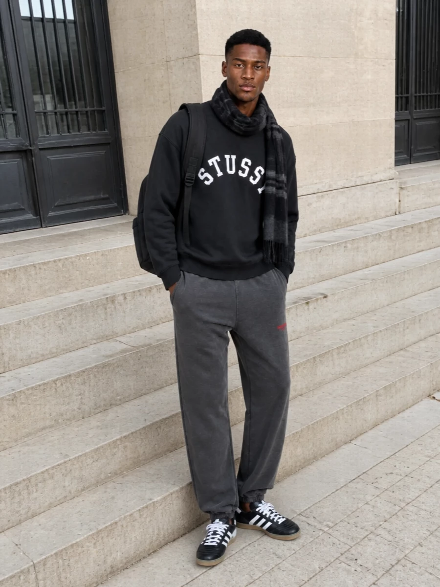 A washed black Stüssy crew sits over charcoal joggers for a head-to-toe street set that feels broken-in but still sharp. Black and white terrace sneakers pick up the logo hit and keep the line low and clean, while the matte black Y-3 pack and grey check scarf add just enough texture for city days and late-night runs.