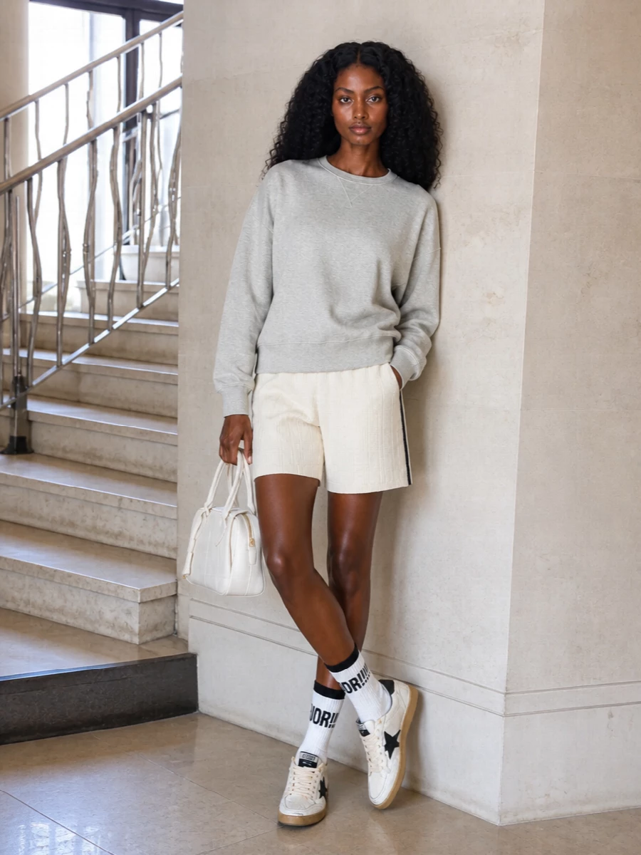 A cream knit Bermuda short meets a slouchy heather-grey sweatshirt for a soft, athletic set that feels ready for coffee runs, flights or city walks. Cream sneakers with black detailing and a structured chalky-white mini bowler keep everything tight, tonal and street focused, while graphic sport socks add a knowing, logo-driven twist.