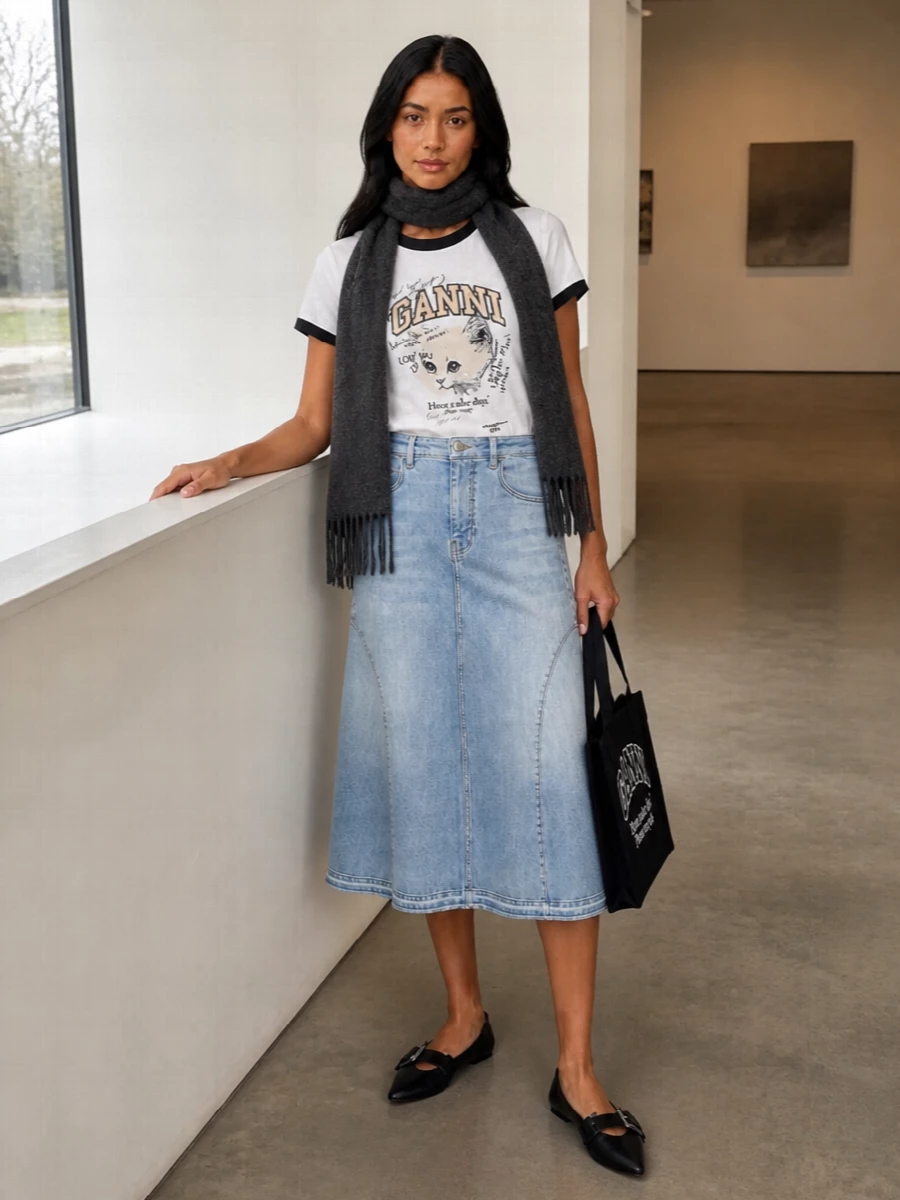 Graphic collegiate energy meets soft volume with the kitten tee tucked into the sculpted light-wash denim midi. Pointed black flats and a matching logo tote pick up the tee’s contrast trims, keeping everything sharp yet easy for day. A simple charcoal scarf adds just enough winter texture without stealing focus from the playful print.