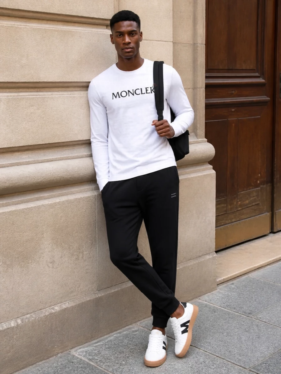 This look leans into Moncler’s clean sport-luxe DNA: the white logo long-sleeve tee sits over sharp black tapered joggers for a streamlined, athletic silhouette. White court sneakers with black accents echo the tee’s palette and keep the outfit polished, while the understated black Moncler backpack finishes things with functional, city-ready ease. It’s a refined off-duty uniform that works from airport lounge to weekend in the city without ever feeling over-styled.