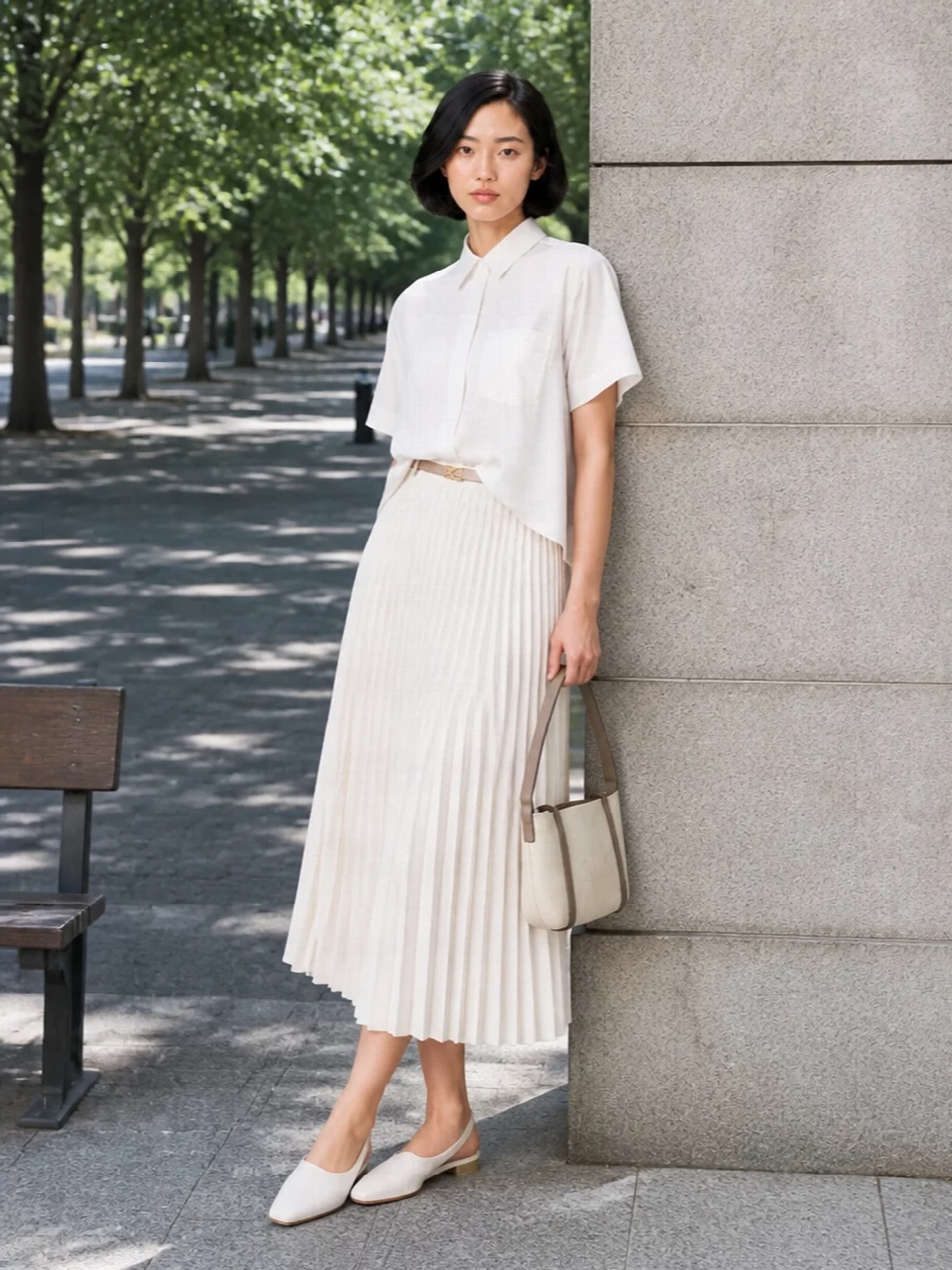 A precise white Sacai shirt tops the pleated ivory skirt, creating a clean, architectural column in soft neutrals. Flat white slingbacks and a compact ivory shoulder bag keep the energy light and purposeful, while a slim beige belt quietly defines the waist and nods to the skirt’s utility hardware. The result is a polished, modern uniform that moves easily from daytime meetings to late-afternoon gallery visits.