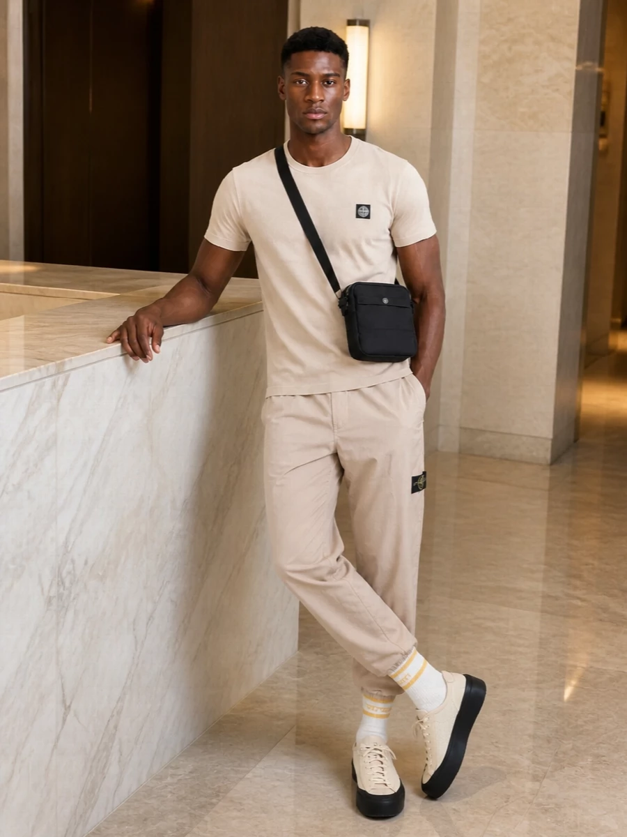 Stone Island’s sand tee and light beige track pants create a clean, tonal base that leans into technical streetwear without feeling heavy. Cream and sand sneakers with a black sole echo the tee’s badge and keep the line sleek, while a compact black crossbody and striped sport socks add just enough utility detail to feel intentional, not busy.