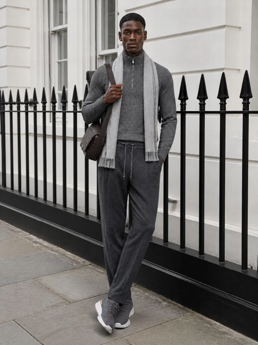 Mid-grey knit and charcoal pleated drawstring trousers create a quietly composed, tonal foundation that feels polished yet relaxed. Charcoal knit runners keep the mood soft and modern rather than sharply dressy, while the espresso intrecciato crossbody and pale grey scarf add discreet richness and texture without disturbing the calm palette.