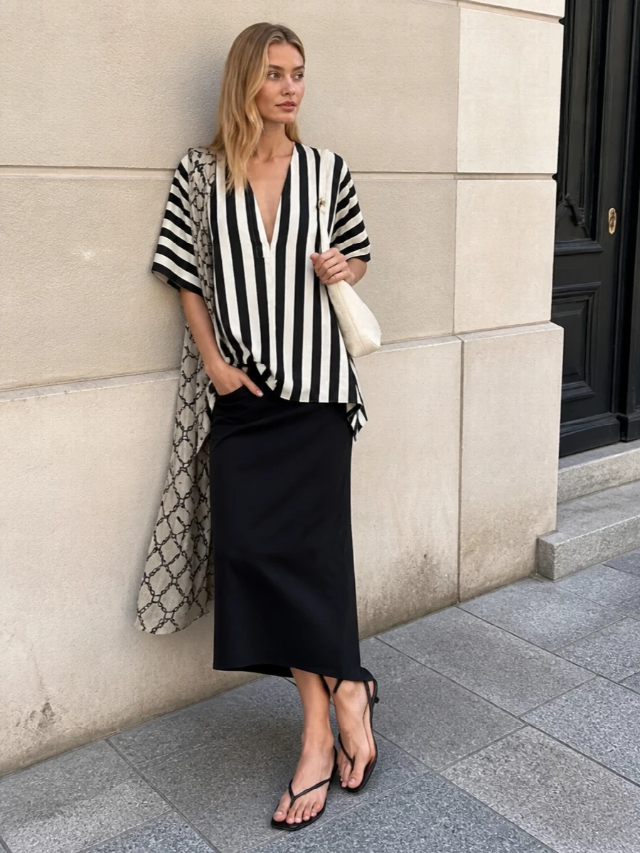 The striped Jacquemus tunic falls over a high-waisted black draped midi skirt, elongating the body and letting the asymmetry do the talking while the lower half stays sleek and sculpted. Bare, graphic lines continue with black thong kitten-heel sandals and a compact off-white shoulder bag, keeping the palette sharply monochrome and the mood quietly architectural. The result is a summer city look that feels directional yet effortless, equally at home from gallery afternoons to late dinners by the sea.