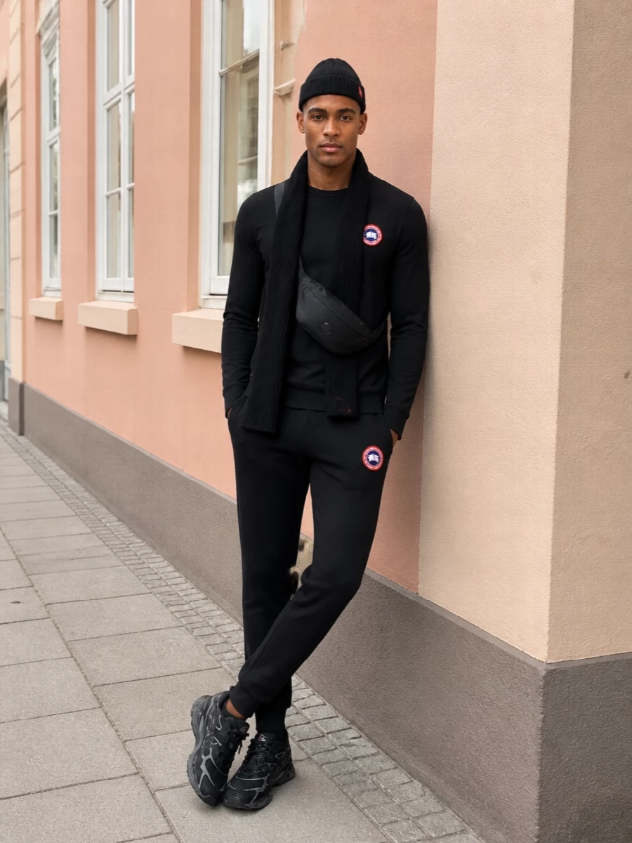 Head-to-toe Canada Goose black anchors this with a quietly athletic mood, the jogger matching the tee’s crest and keeping the silhouette clean but relaxed. Technical Asics runners add a sculpted base that still reads minimal, while a low-profile Moncler belt bag and ribbed beanie-scarf set keep hands free and the red accents feeling deliberate rather than loud.