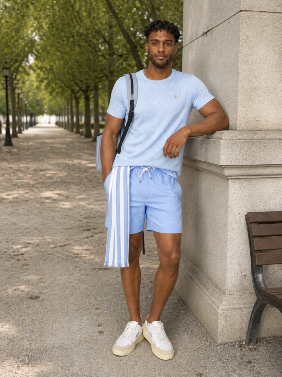 Soft blues and clean lines keep this outfit firmly in that quiet Polo universe, with the tee and washed deck shorts reading as an easy tonal set. Neutral court sneakers and a streamlined steel blue backpack modernise the prep, while a striped scarf adds a subtle coastal accent without breaking the calm palette. It is an off-duty summer uniform that feels relaxed but still considered from coffee run to beachside drinks.