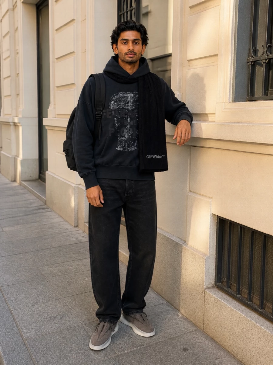 Washed black on washed black keeps the Fear Of God hoodie grounded, with the straight-leg trouser giving just enough structure to the relaxed silhouette. Charcoal taupe runners soften the edge at the floor, while a clean black backpack and discreet logo scarf finish the city-ready uniform without diluting the graphic attitude up top.