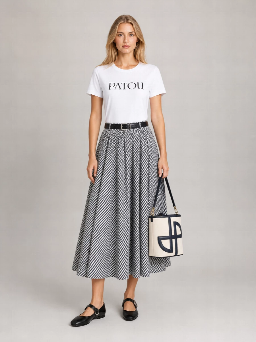A crisp white logo tee tops the striped Patou midi, keeping the whole silhouette light, graphic and easy for warm city days. Black Mary Jane flats and a structured monogram bucket bag ground the volume with Parisian neatness rather than weight. A slim black belt ties everything together, accenting the waist so the skirt can move with that soft, swishy drama.
