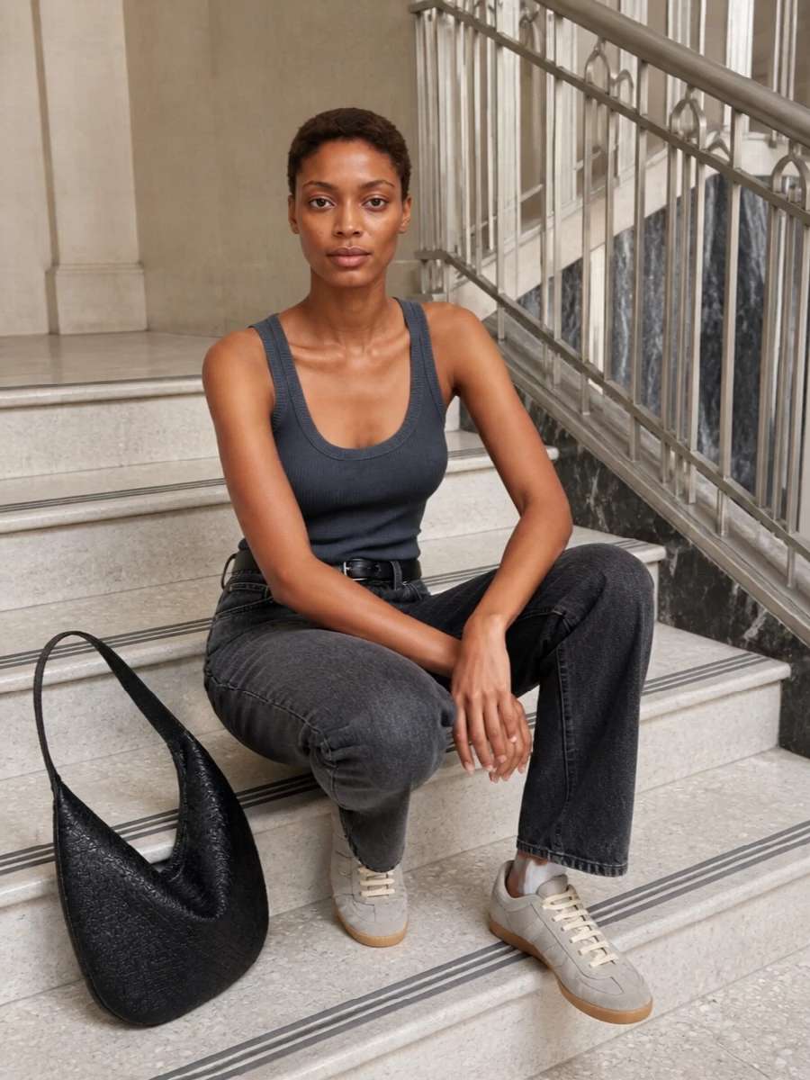 A charcoal-on-charcoal palette keeps this pairing of high-rise Haikure denim and a ribbed tank quietly intentional, reading as a clean column from shoulder to hem. Soft grey sneakers and a sculptural black shoulder bag keep the mood relaxed but precise, while a slim black belt subtly defines the waist without breaking the minimal line.