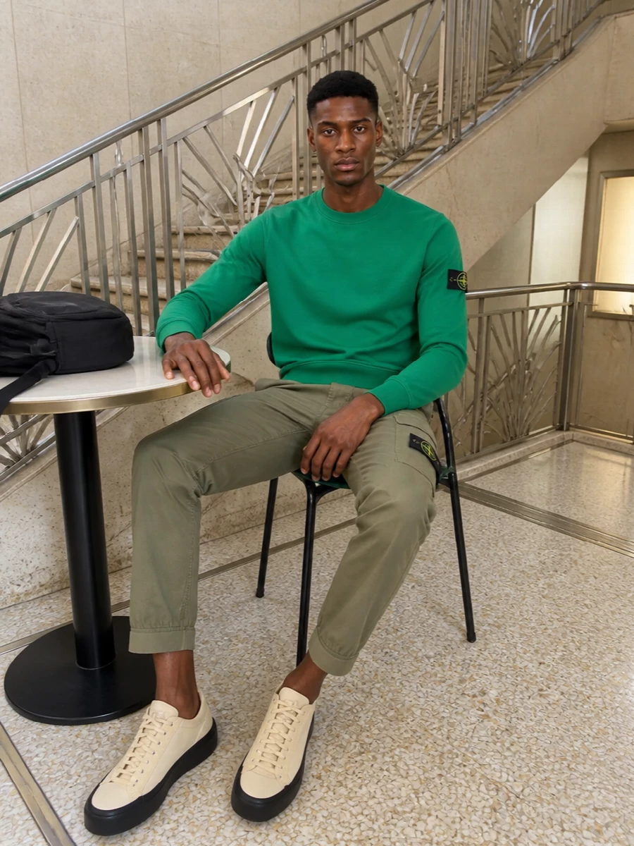 Vivid green on top meets muted olive below for a clean Stone Island take on sport-street that feels sharp without trying too hard. Neutral technical sneakers and a black backpack keep the palette grounded and functional, so the colour hit stays focused on the crewneck and badge details.